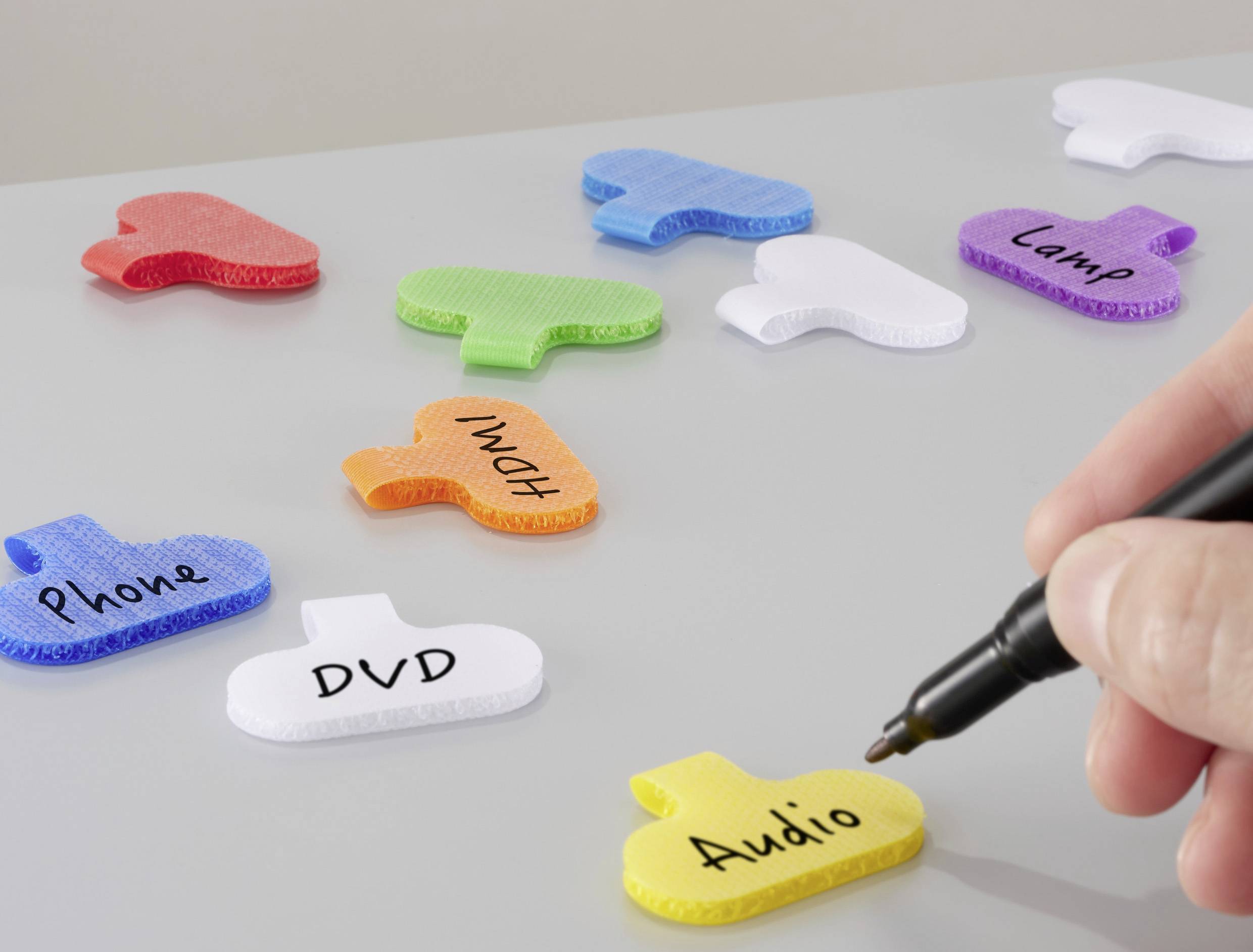 Een hand schrijft op een gele sleutelhanger met het label 'Audio'; verschillende andere gelabelde tags zoals 'Telefoon', 'DVD', 'Lamp' zijn verspreid over een oppervlak.