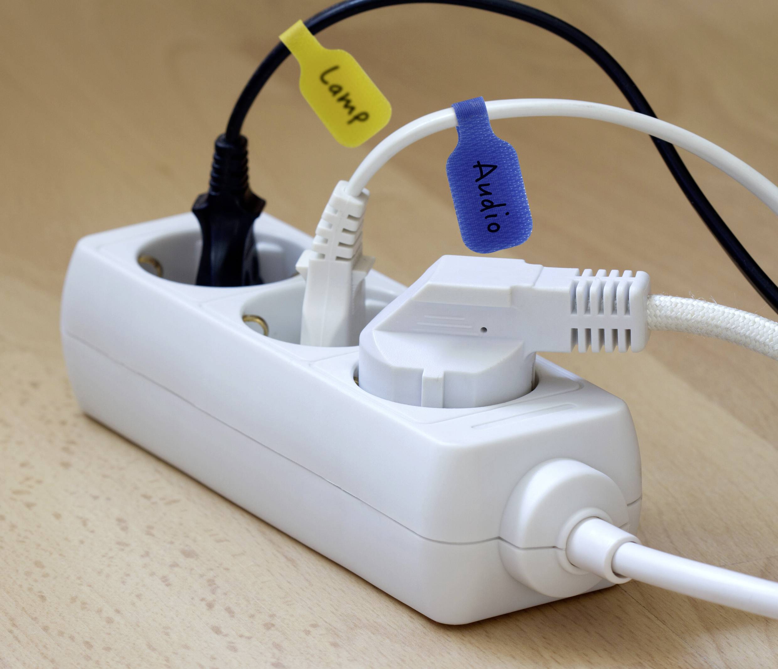 Een witte stekkerdoos op een houten oppervlak met verschillende snoeren aangesloten, voorzien van een gele tag met het label 'Lamp' en een blauwe tag met het label 'Audio'.