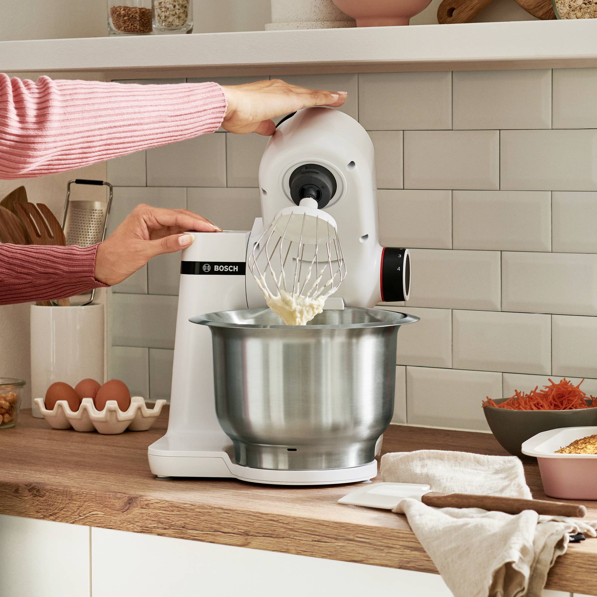 Een persoon gebruikt een witte standmixer met een garde-opzetstuk om ingrediënten in een kom te mengen, omringd door een keukenopstelling met eieren en specerijevenbakjes.