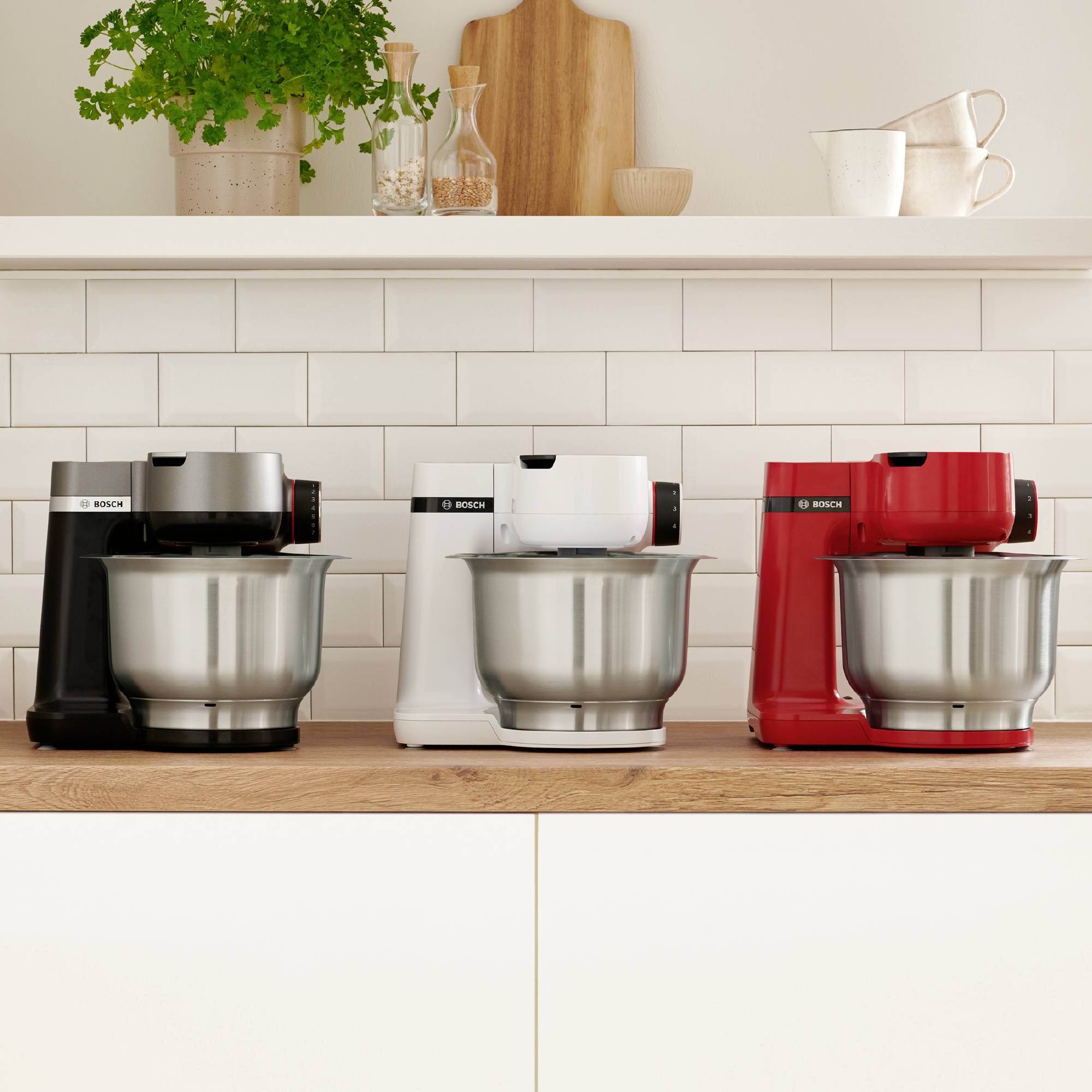 Drie standmixers in zwart, wit en rood op een aanrecht onder een plank met planten en kopjes tegen een wit betegelde muur.