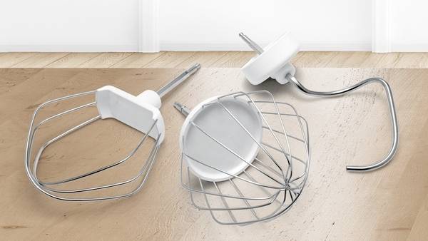 Drie roestvrijstalen menghaakjes, waaronder een garde, deeghaak en platte mengspatel, liggen op een houten ondergrond.