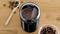 Een zwarte koffiemolen gevuld met gemalen koffie op een houten oppervlak, omringd door verspreid liggende koffiebonen en een houten lepel.