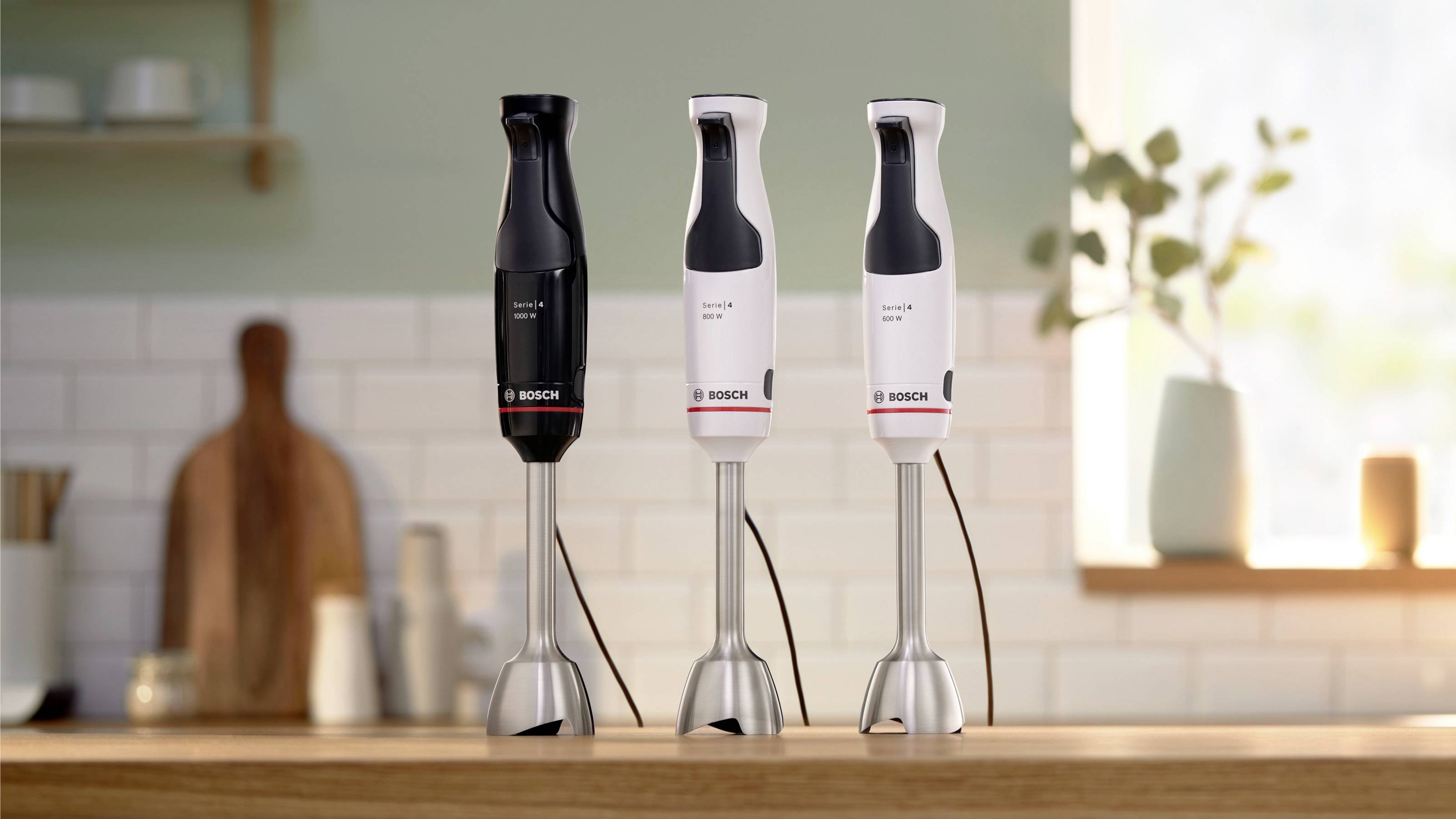 Drie Bosch staafmixers in zwart en wit worden tentoongesteld op een aanrecht, met een wazig keukendecor op de achtergrond.