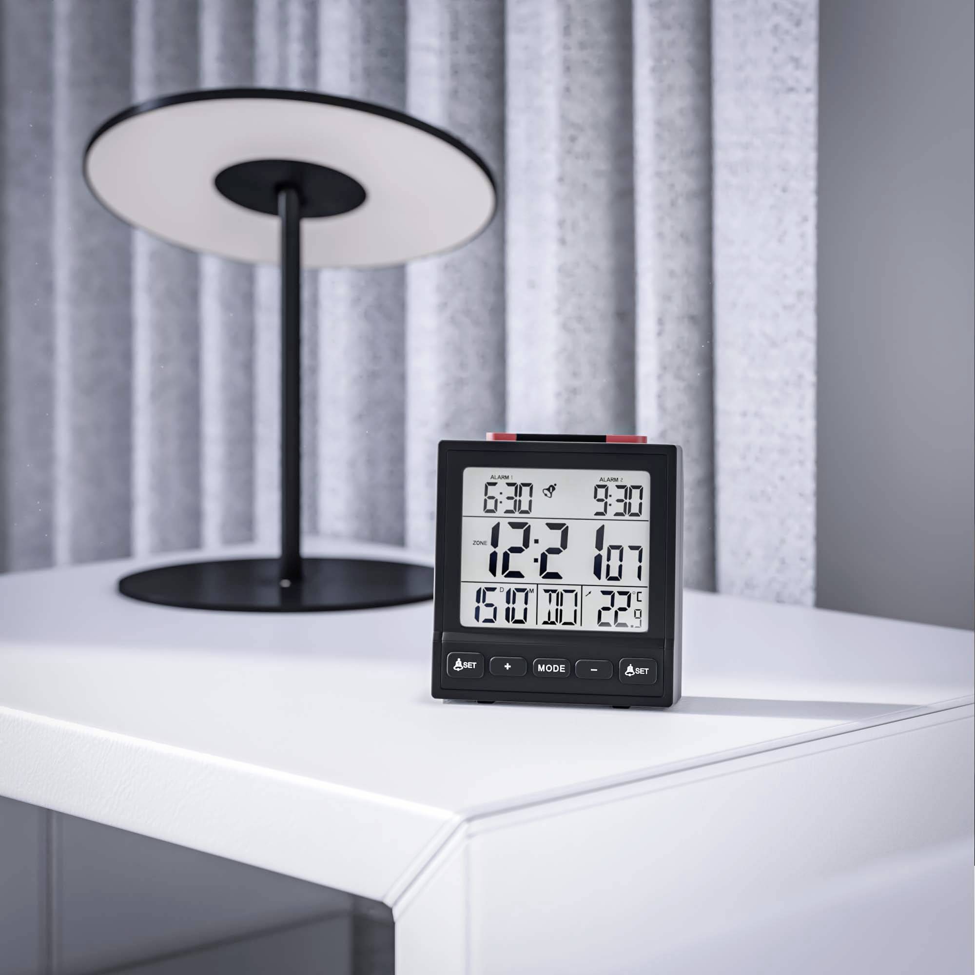 Een digitale klok die '6:30' aangeeft met temperatuurmetingen staat op een witte tafel naast een moderne, ronde lamp.