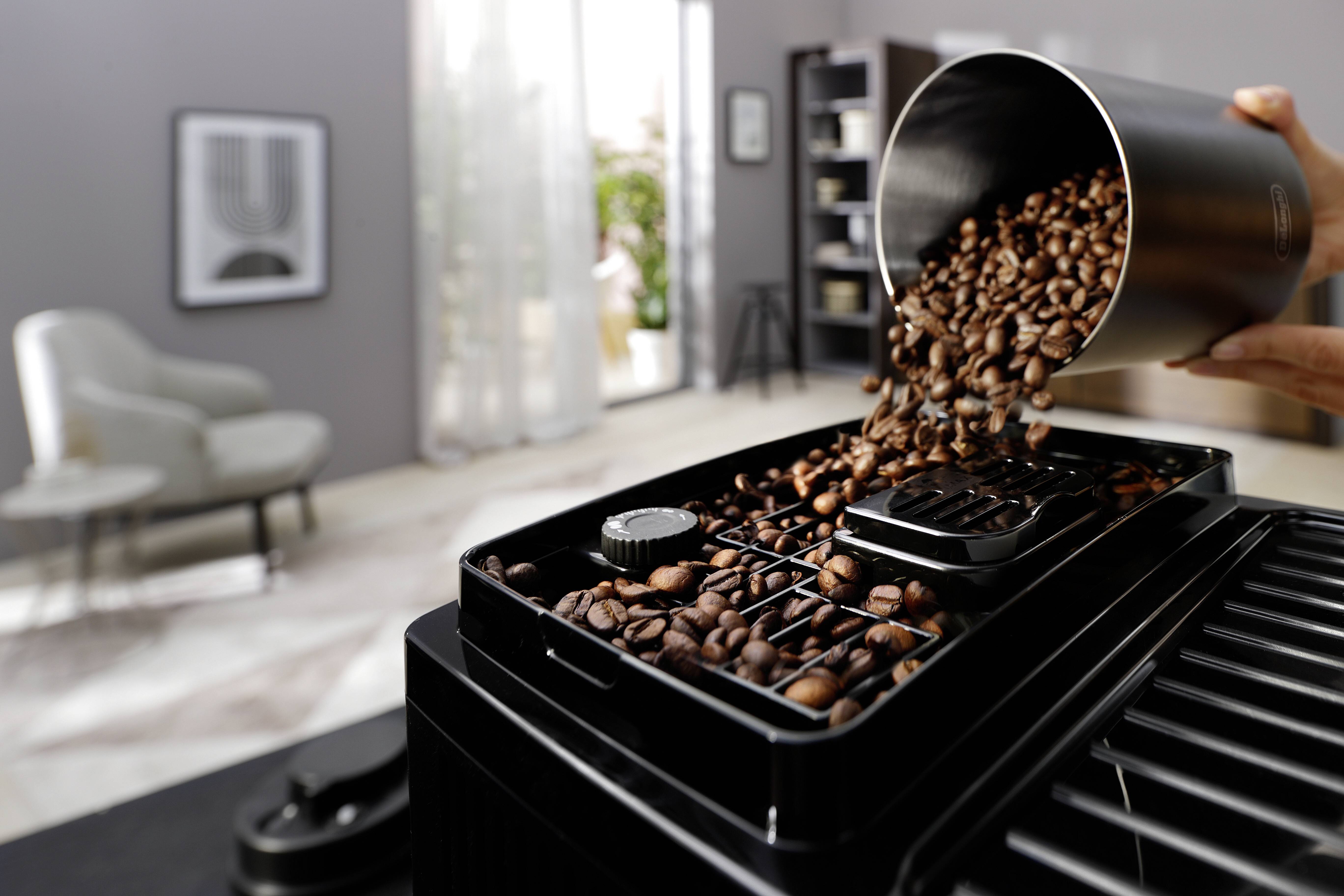 Koffiebonen worden in de trechter van een zwarte koffiemolen gegoten in een moderne, helder verlichte ruimte met zachte meubels op de achtergrond.