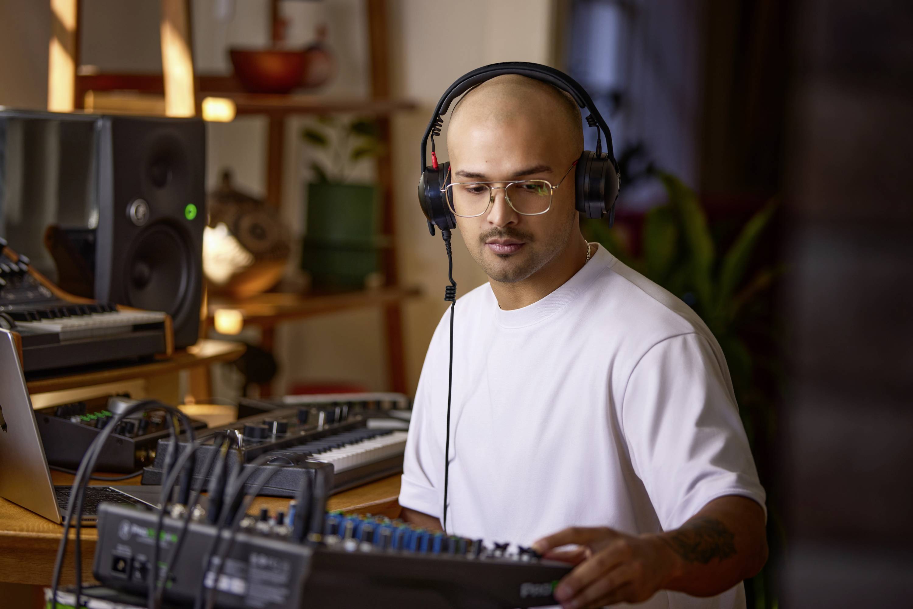 Een persoon met koptelefoon en een wit shirt bedient geluidsapparatuur in een muziekstudio, omringd door speakers en synthesizers.