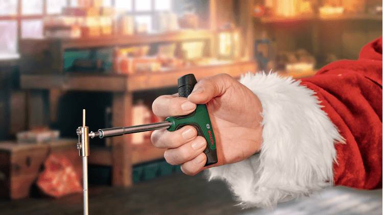 Een hand in een kerstmanpak houdt een groene en zwarte schroevendraaier binnen vast, wat wijst op assemblage of vakmanschap met een feestelijk thema.