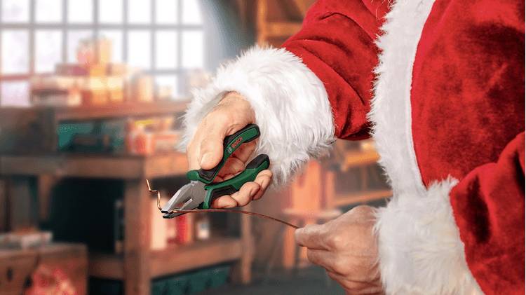 Een persoon in een kerstmanpak gebruikt een tang om draad door te knippen in een werkplaats vol gereedschappen en dozen.