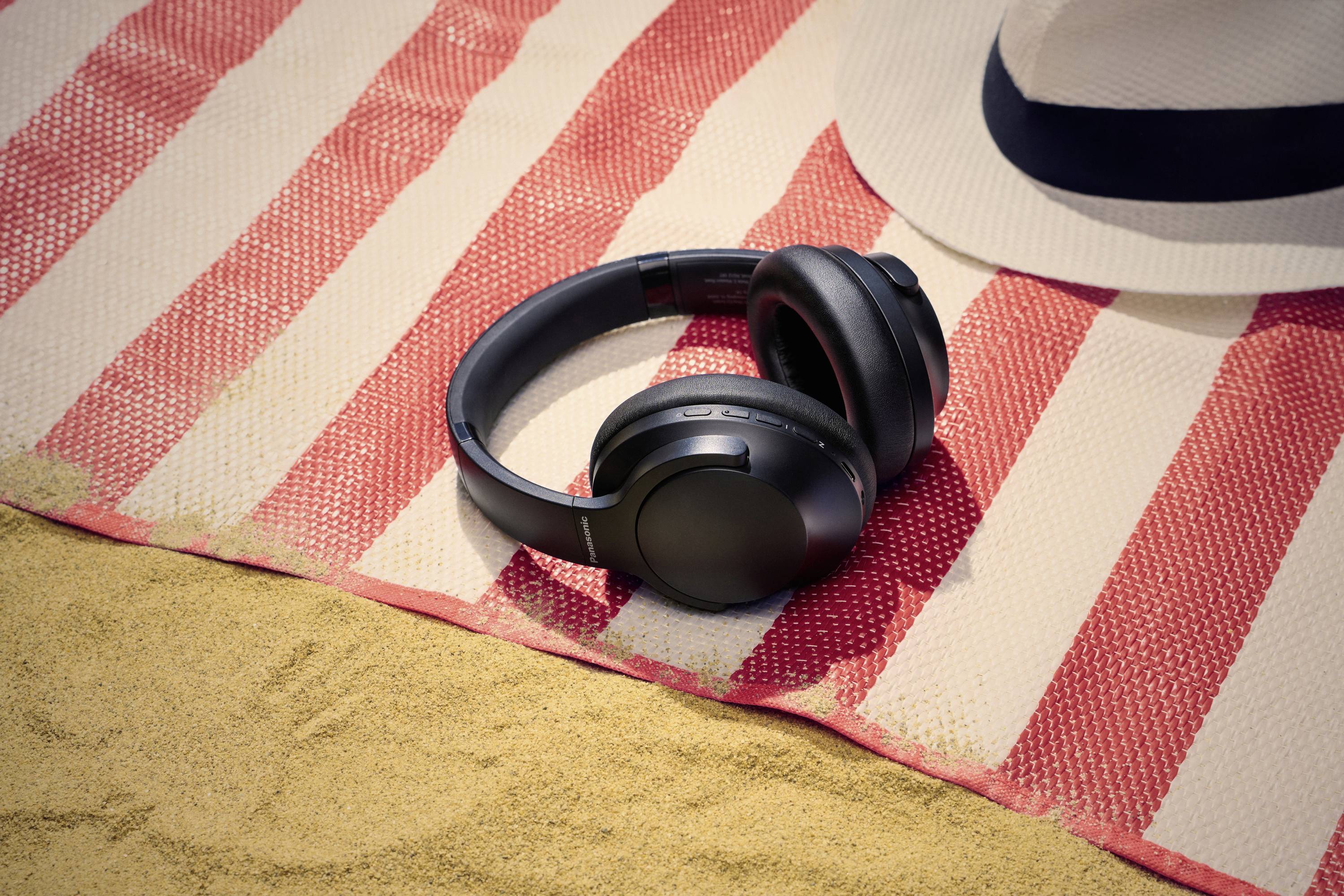 Koptelefoon die rust op een gestreepte strandmat naast een zonnehoed op zandgrond, wat een ontspannende strandopstelling met muziek suggereert.