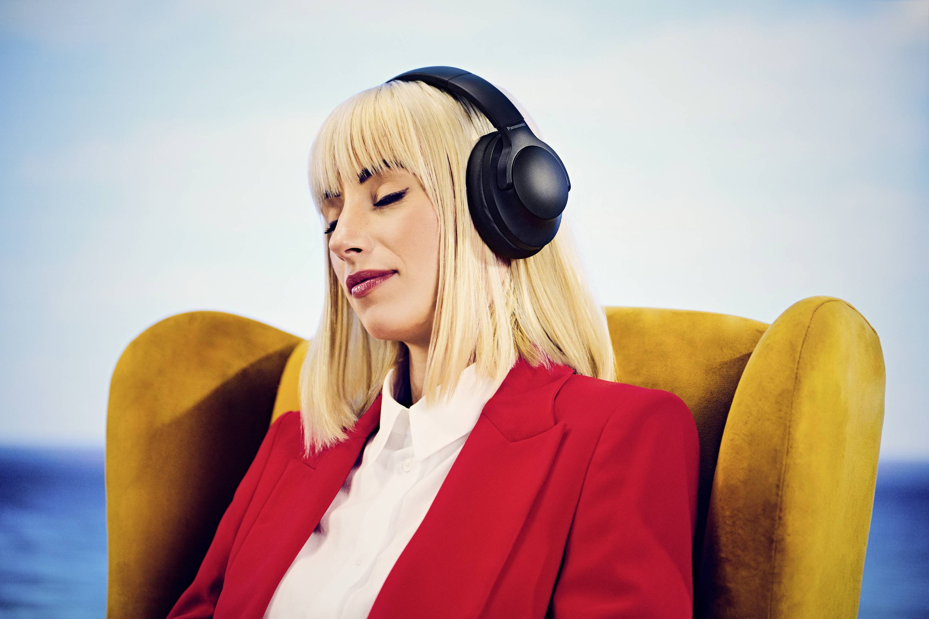 Een persoon met koptelefoon zit ontspannen in een gele stoel, ogen gesloten, tegen een onscherpe blauwe achtergrond die kalmte of muziekgenot suggereert.