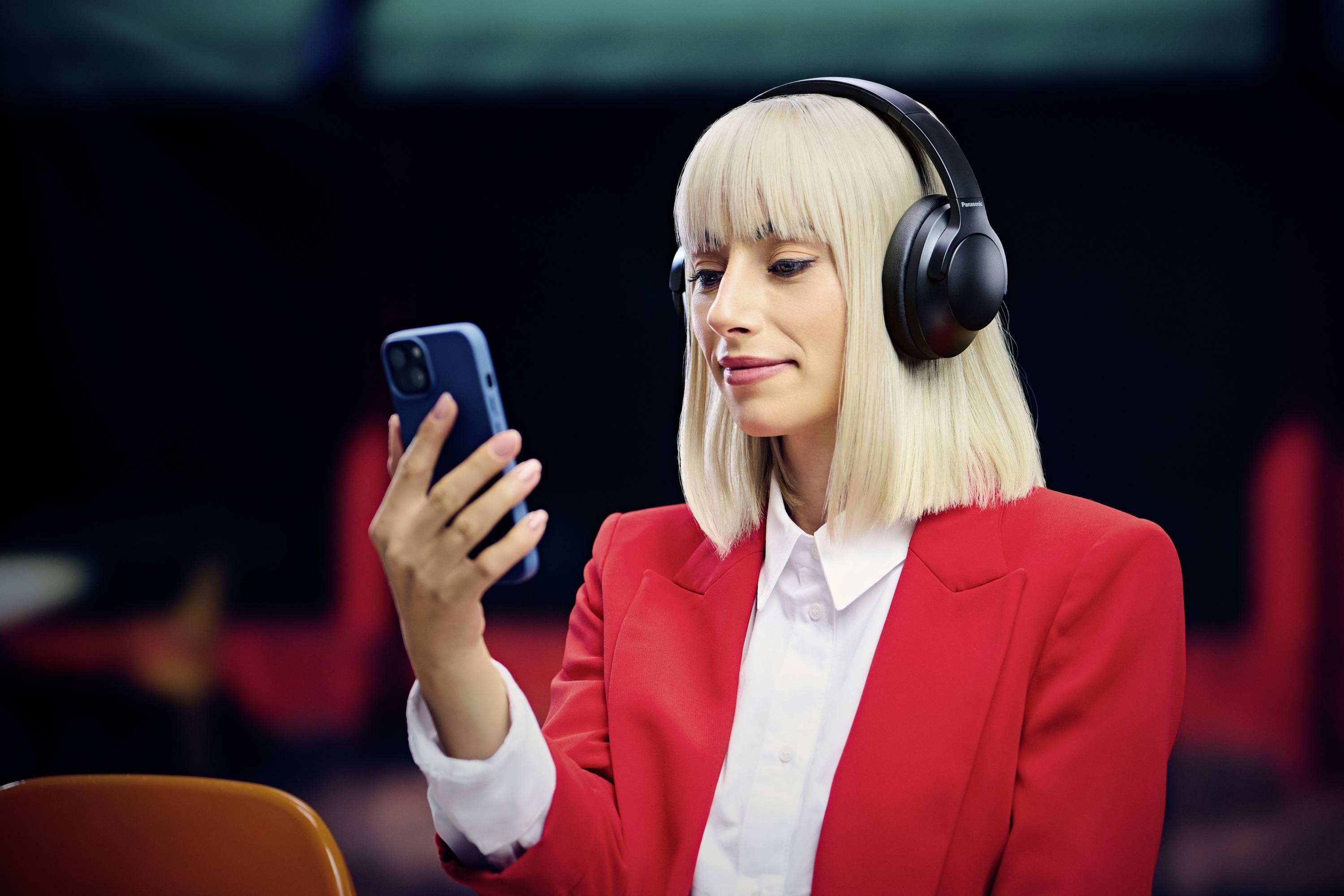 Een persoon met koptelefoon aan en een rode blazer kijkt naar een smartphone, waarschijnlijk in gesprek met muziek of een podcast binnen.