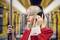 Een persoon met blond haar draagt koptelefoons en een rode jas, staat in de metro terwijl hij/zij zich vasthoudt aan een steun en een geconcentreerde uitdrukking heeft.