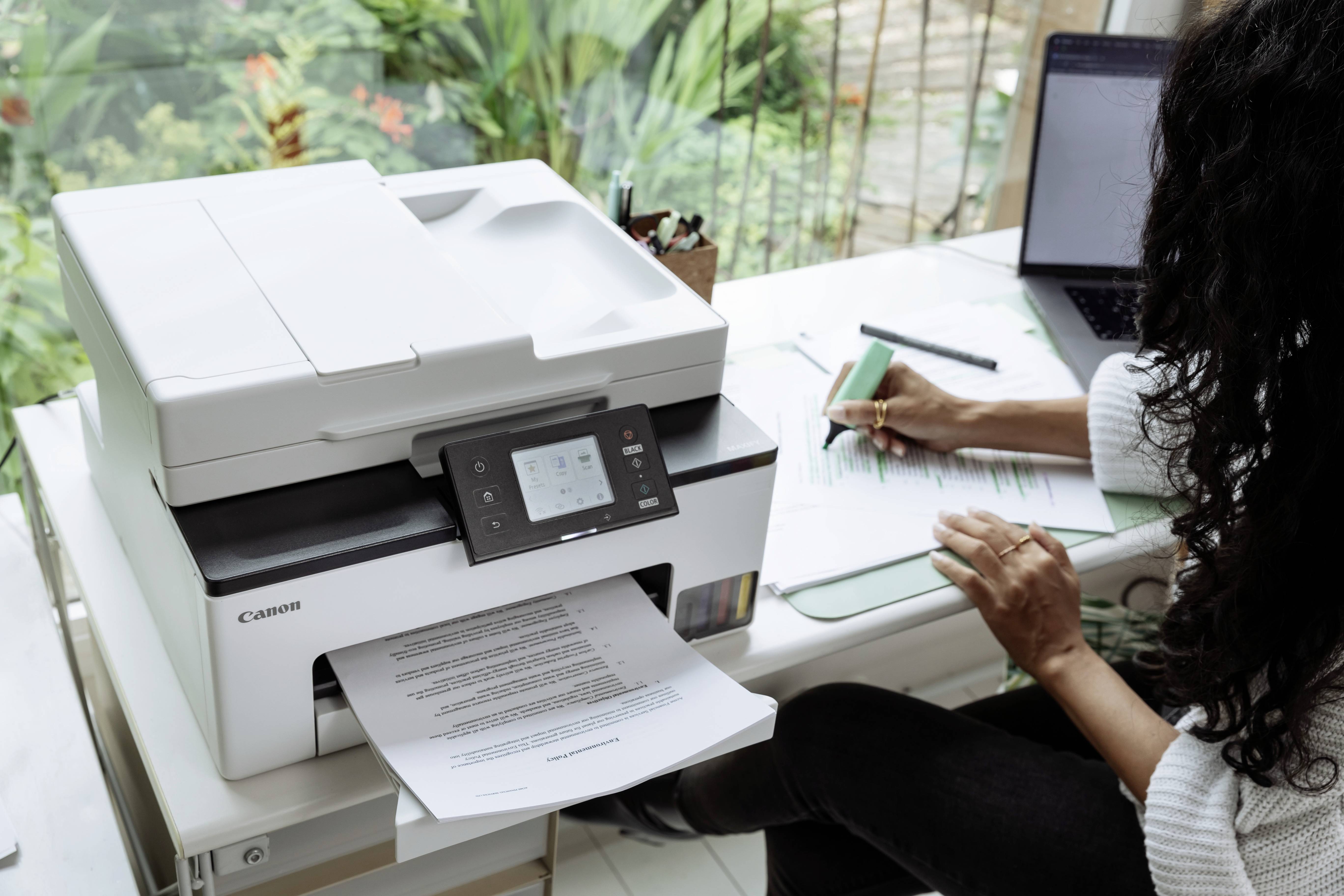 Een persoon zit aan een bureau in een thuiskantoor, met een printer naast een laptop. De printer is actief aan het printen van een document.