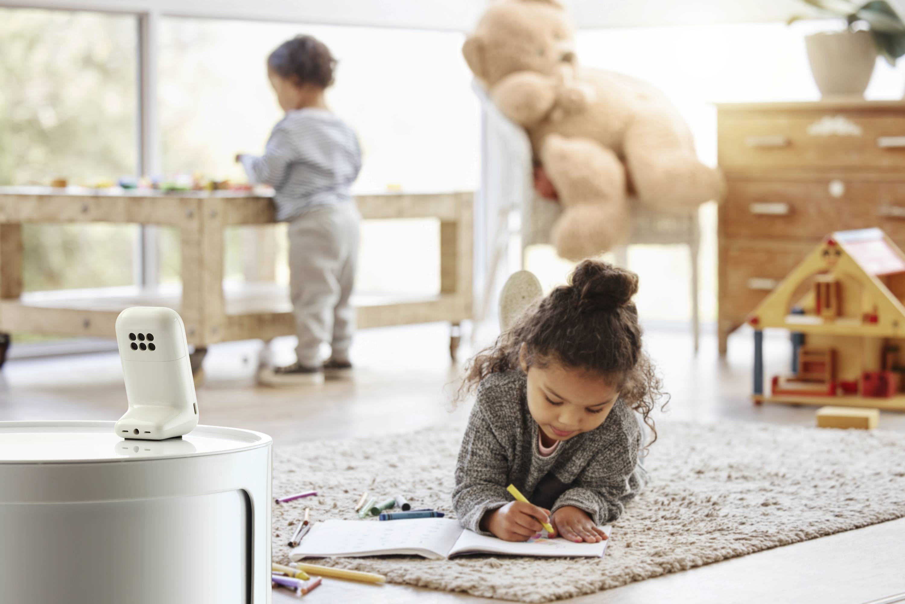Een kind tekent op papier terwijl het op een vloerkleed ligt. Op de achtergrond speelt een ander kind aan een tafel. Een babyfoon is nabij zichtbaar.