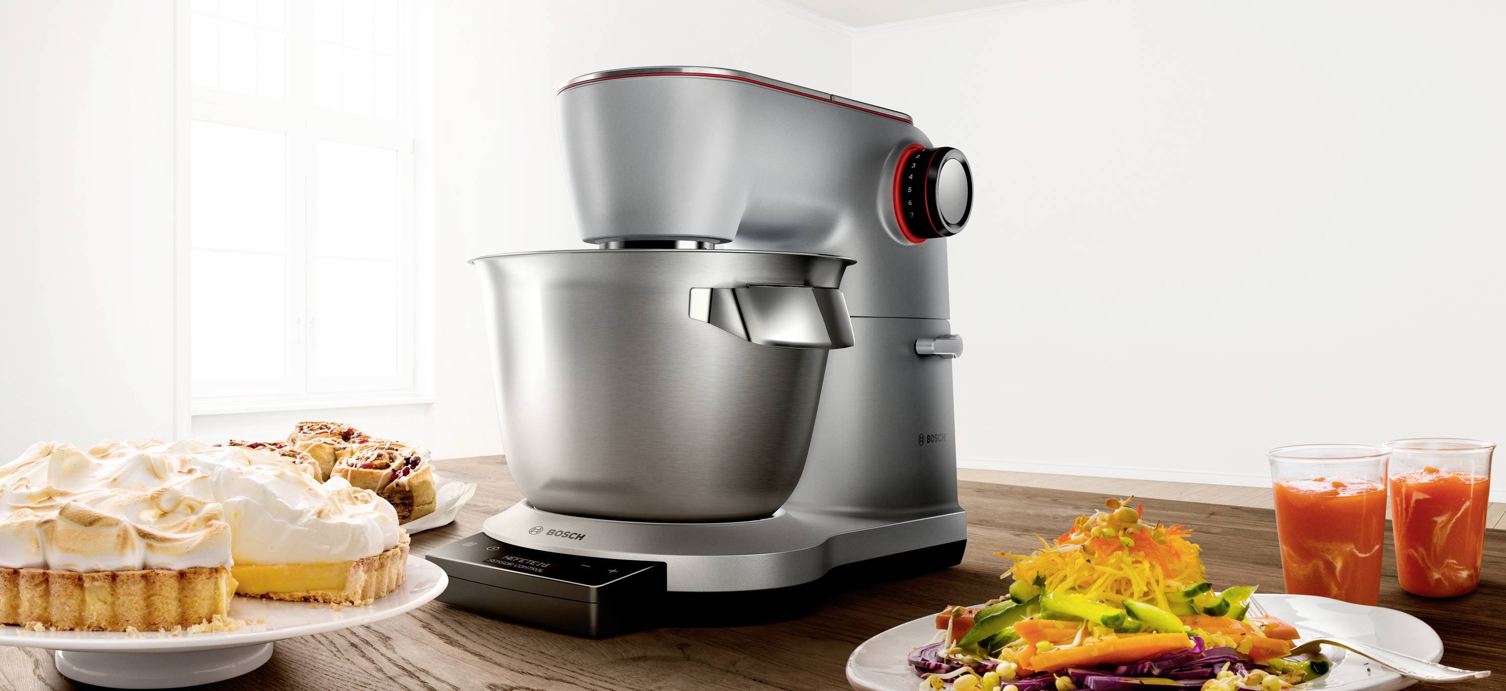 Een zilveren keukenmixer op een houten tafel, omringd door borden met cake, salade en twee glazen sap.