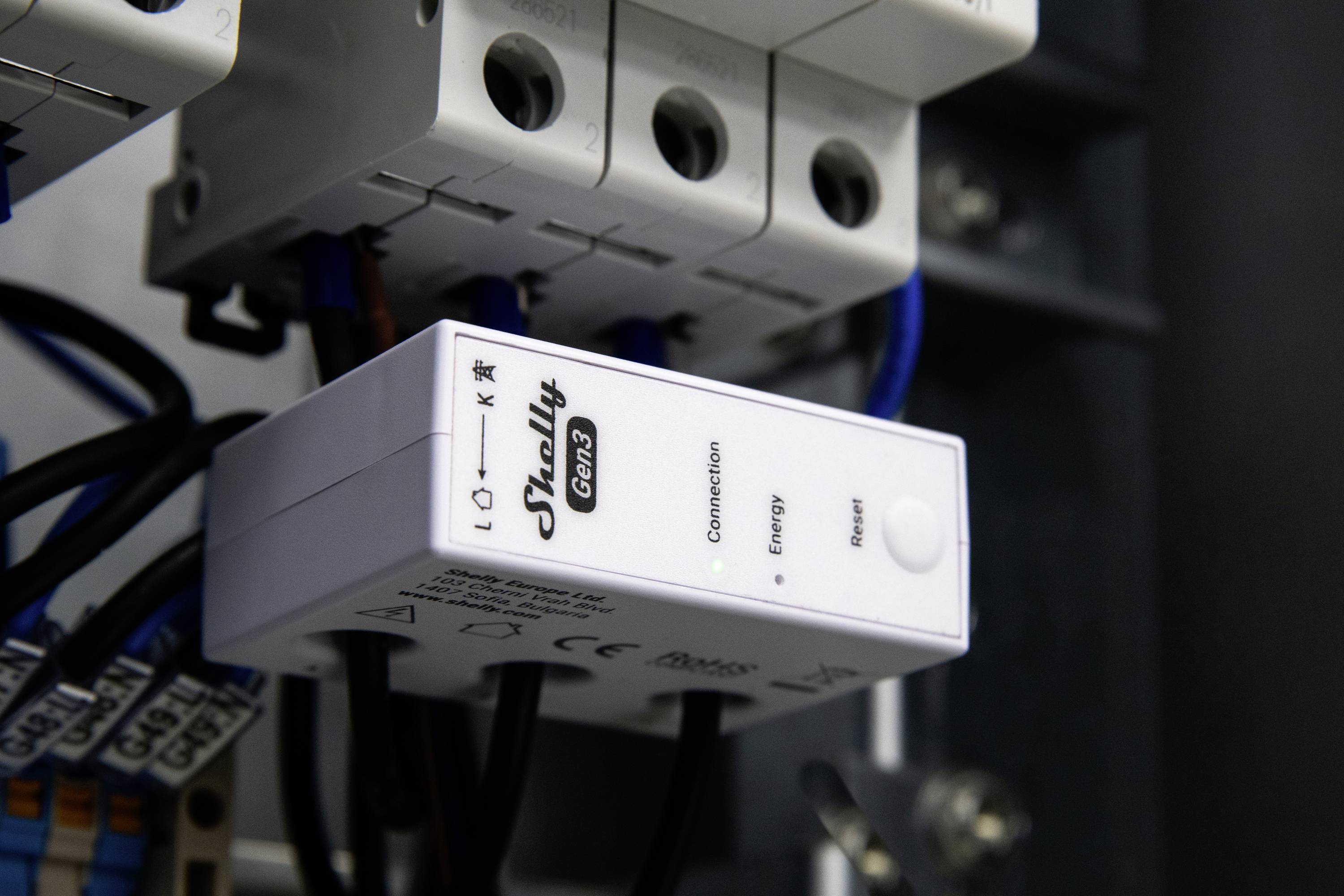 Een witte Shelly Gen3-apparaat met aangesloten en energie-indicatoren, gemonteerd in een elektrisch paneel met zichtbare draden en componenten.