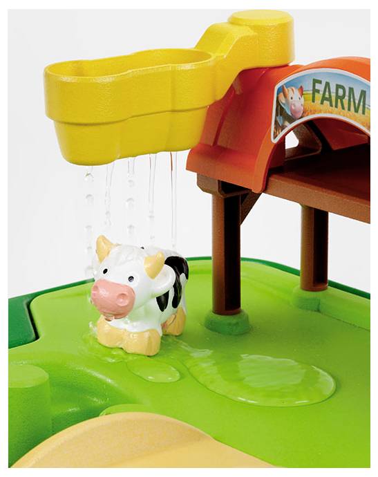 Een speelgoed koe staat onder een gele waterstraal, die water naar beneden laat druppen. Een rode boog met het label 'BOERDERIJ' bevindt zich boven de koe op een groene basis.