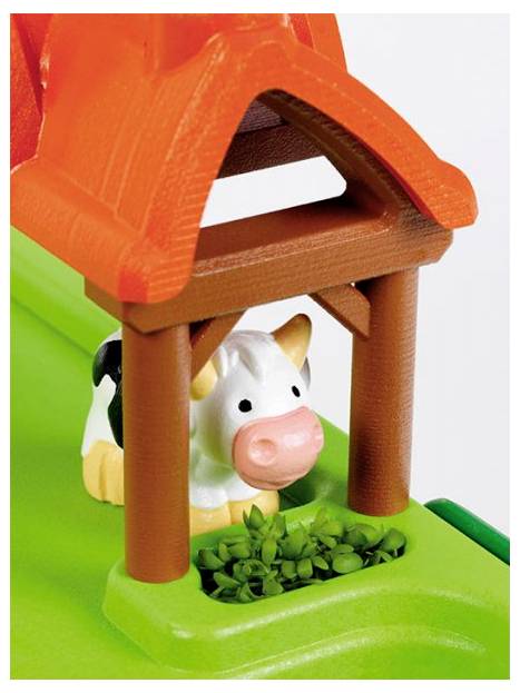 Een speelgoed koe staat onder een speelgoed schuur dak naast een groen platform met kleine planten.