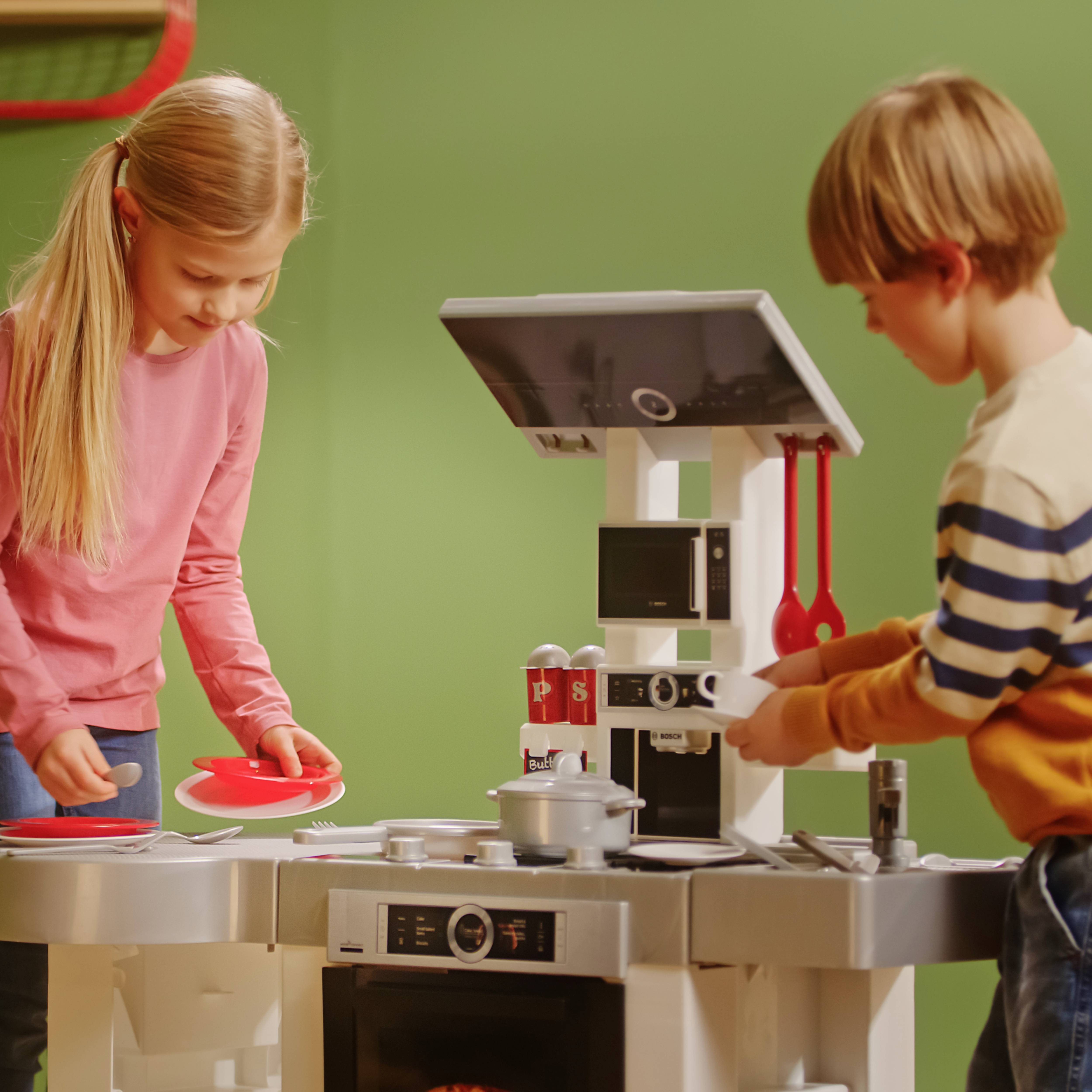 Twee kinderen spelen met een speelkeukenset en doen alsof ze aan het koken zijn. Het meisje houdt een rood bord vast, terwijl de jongen met het fornuis bezig is.