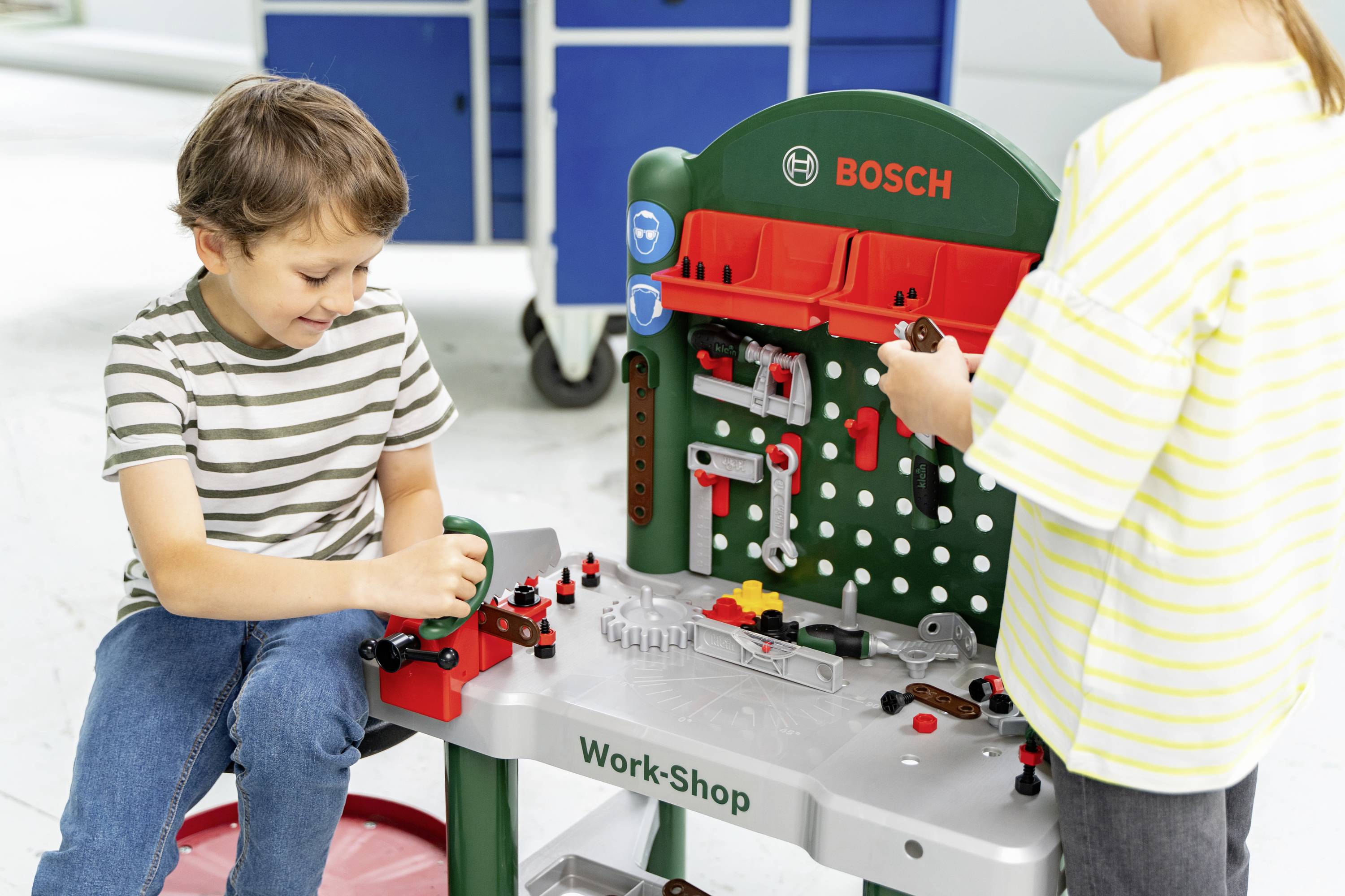 Een jonge jongen en meisje spelen met een Bosch speelgoedwerkplaats, waarbij ze speelgereedschap gebruiken op een werkbank met verschillende accessoires en uitrusting.