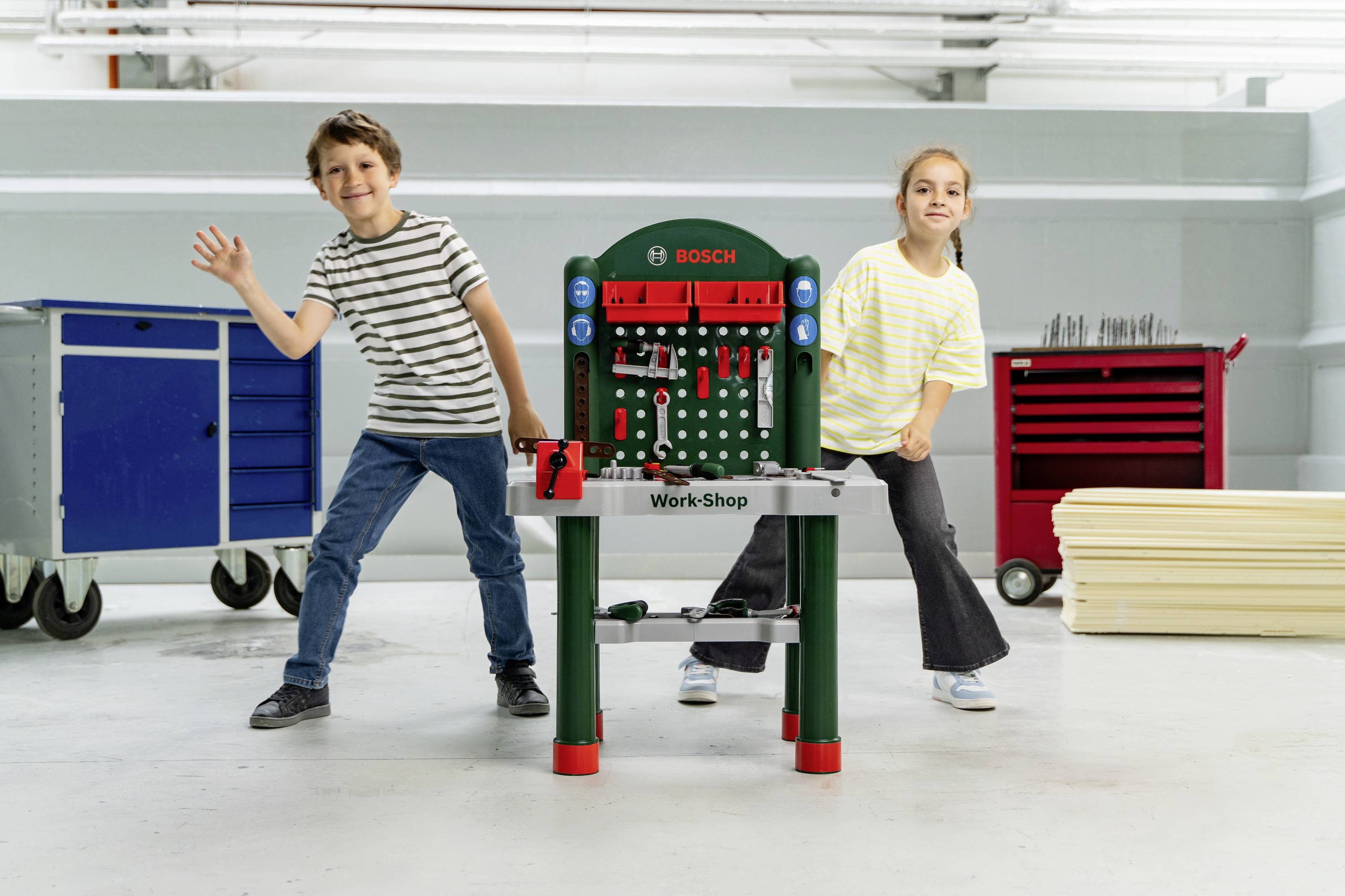 Twee kinderen poseren speels met een houten speelgoedgereedschapskist in een lichte ruimte. De werkbank toont gereedschappen, en twee verrijdbare gereedschapskasten staan op de achtergrond.