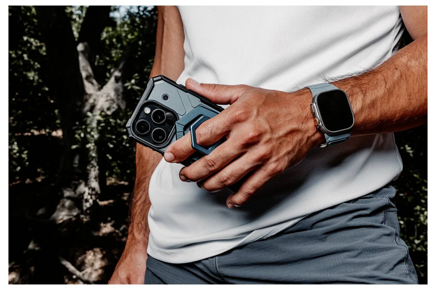 Een persoon draagt een wit shirt en een smartwatch terwijl hij een smartphone met een robuuste beschermhoes vasthoudt in een buitenomgeving met bomen op de achtergrond.