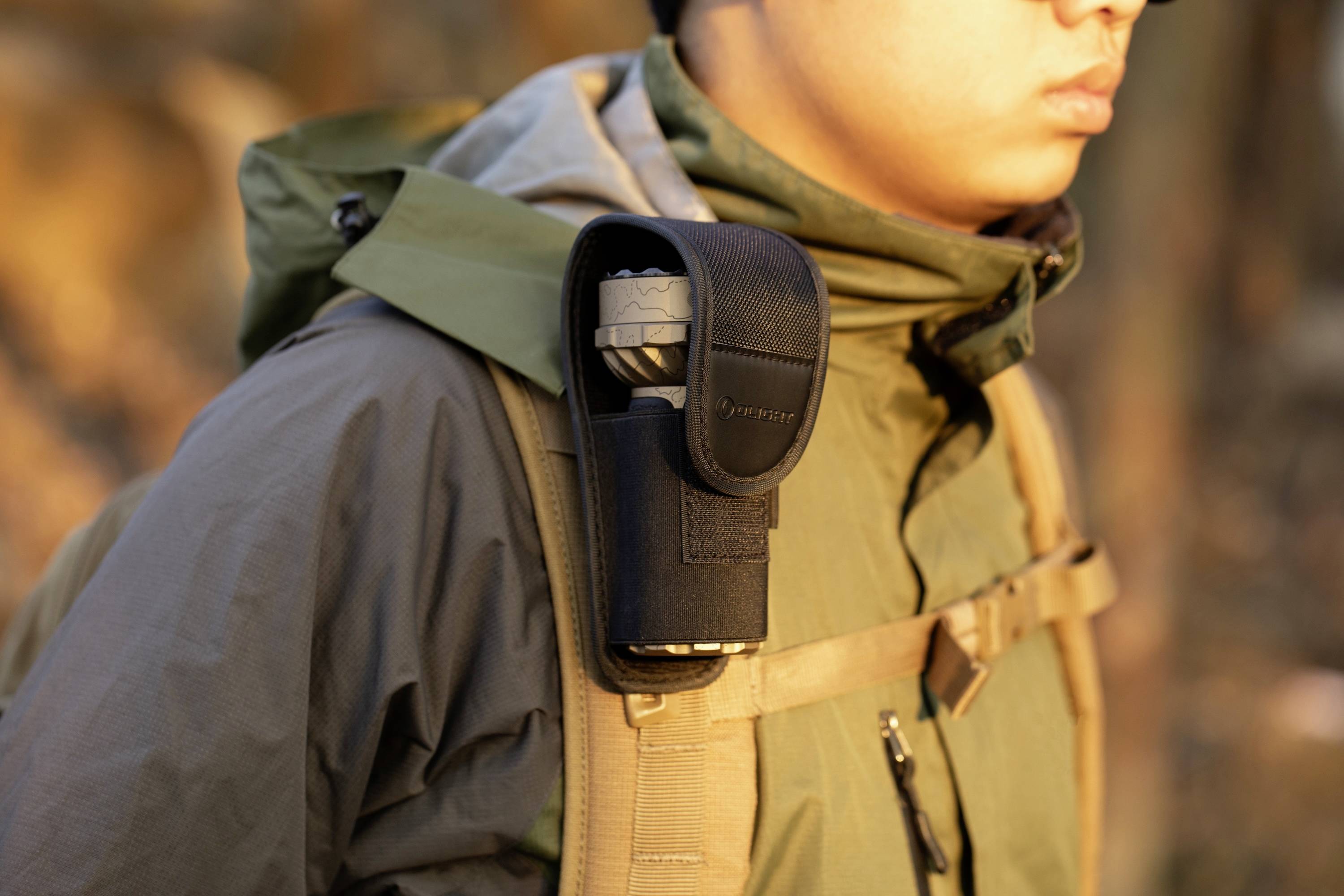 Een persoon in outdoorkleding draagt een opgerolde paraplu in een zwarte houder die aan de draagriem van zijn rugzak is bevestigd, terwijl hij in een natuurlijke omgeving loopt.