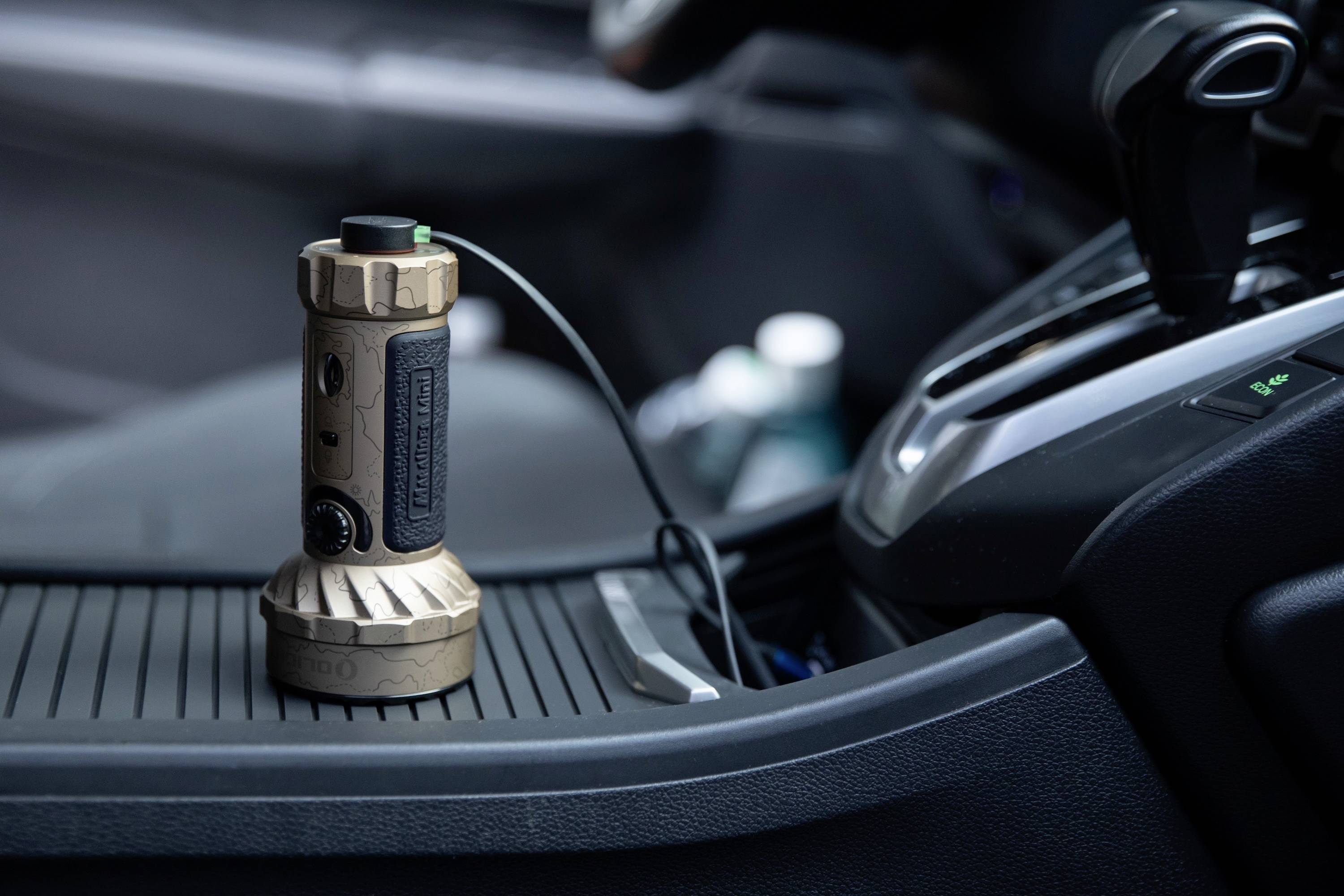 Een cilindrische gadget met een tactische greep wordt geplaatst op de middenconsole van een auto. Hij is verbonden met een kabel, wat elektronische functionaliteit suggereert.