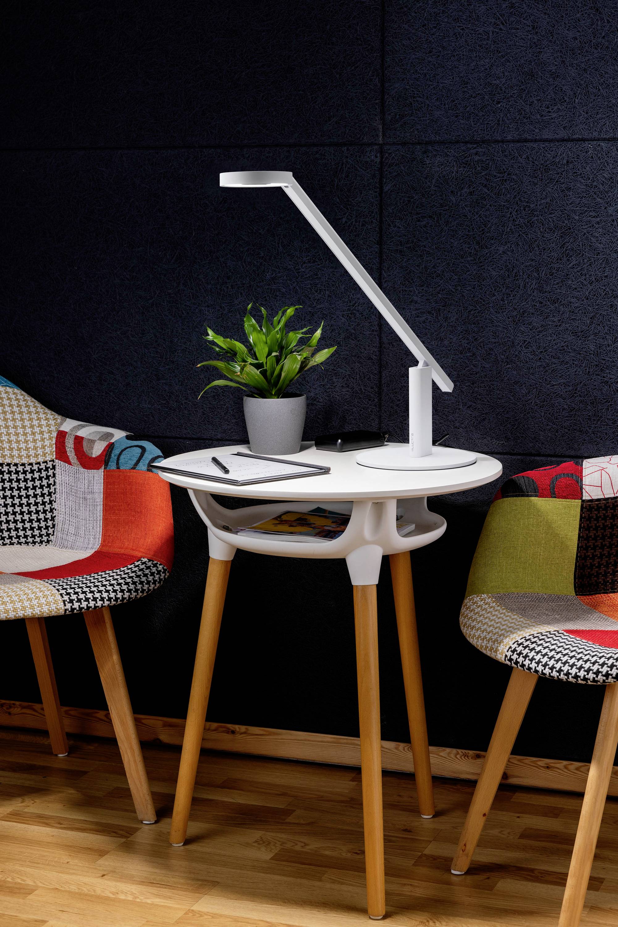 Een kleine witte tafel met een moderne lamp, een potplant en een pen en notitieboek, geflankeerd door twee kleurrijke, gestileerde stoelen.