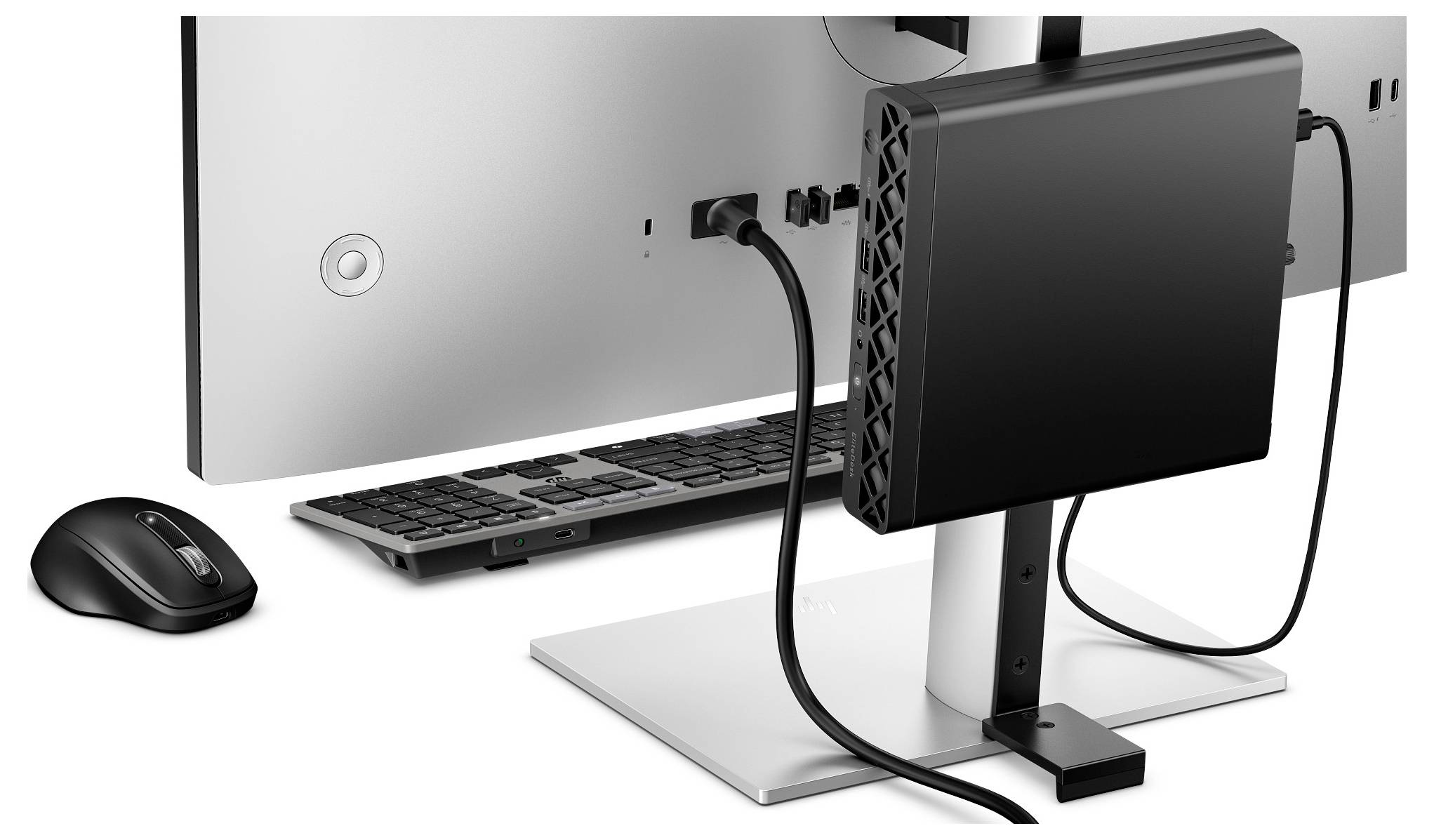 Een dunne pc is gemonteerd aan de achterkant van een monitorstandaard, verbonden met kabels. Een draadloze muis en toetsenbord zijn in de buurt geplaatst.