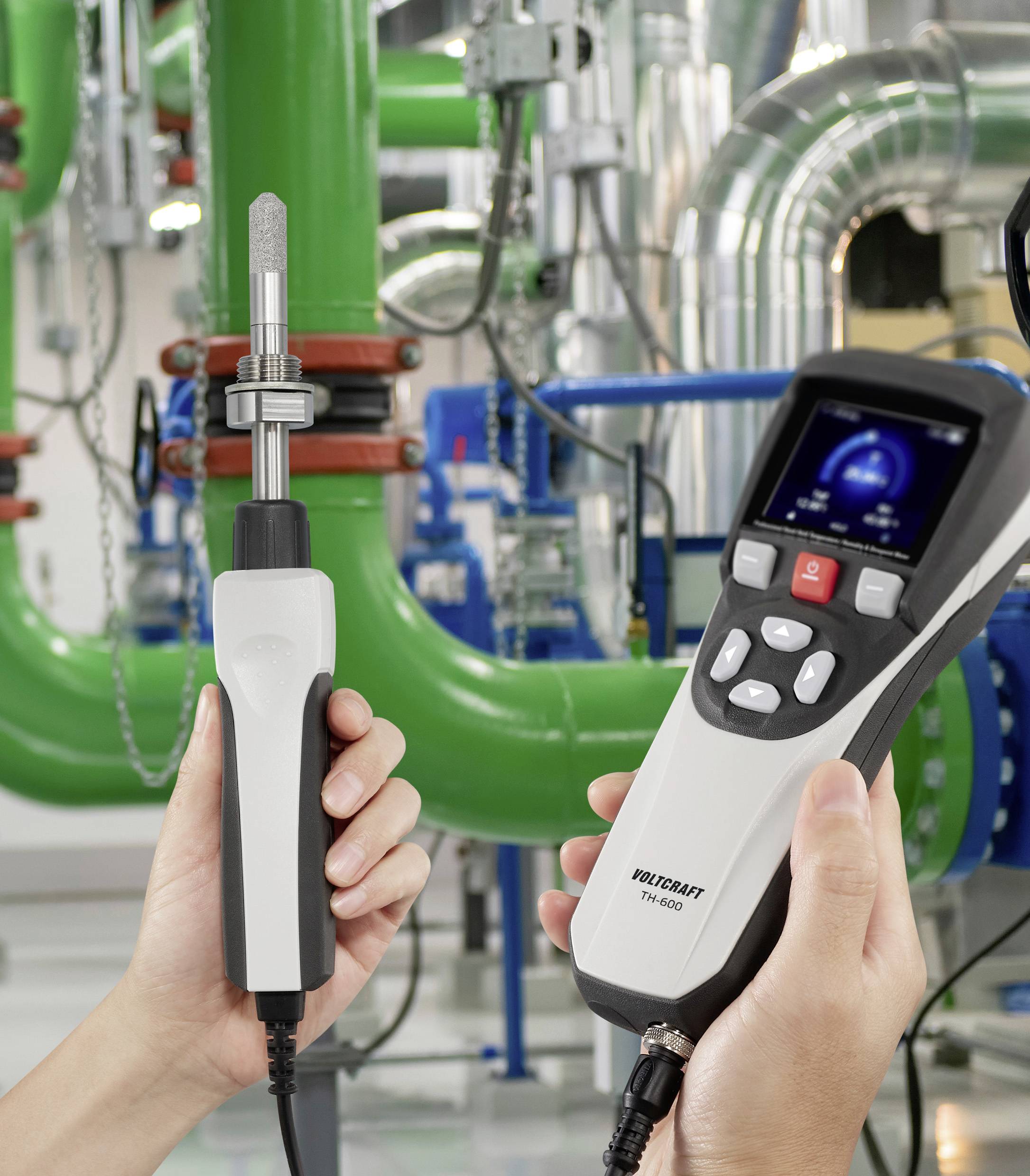 Een persoon houdt een digitale thermometer en sensor vast in een industriële faciliteit met groene buizen en machines op de achtergrond.