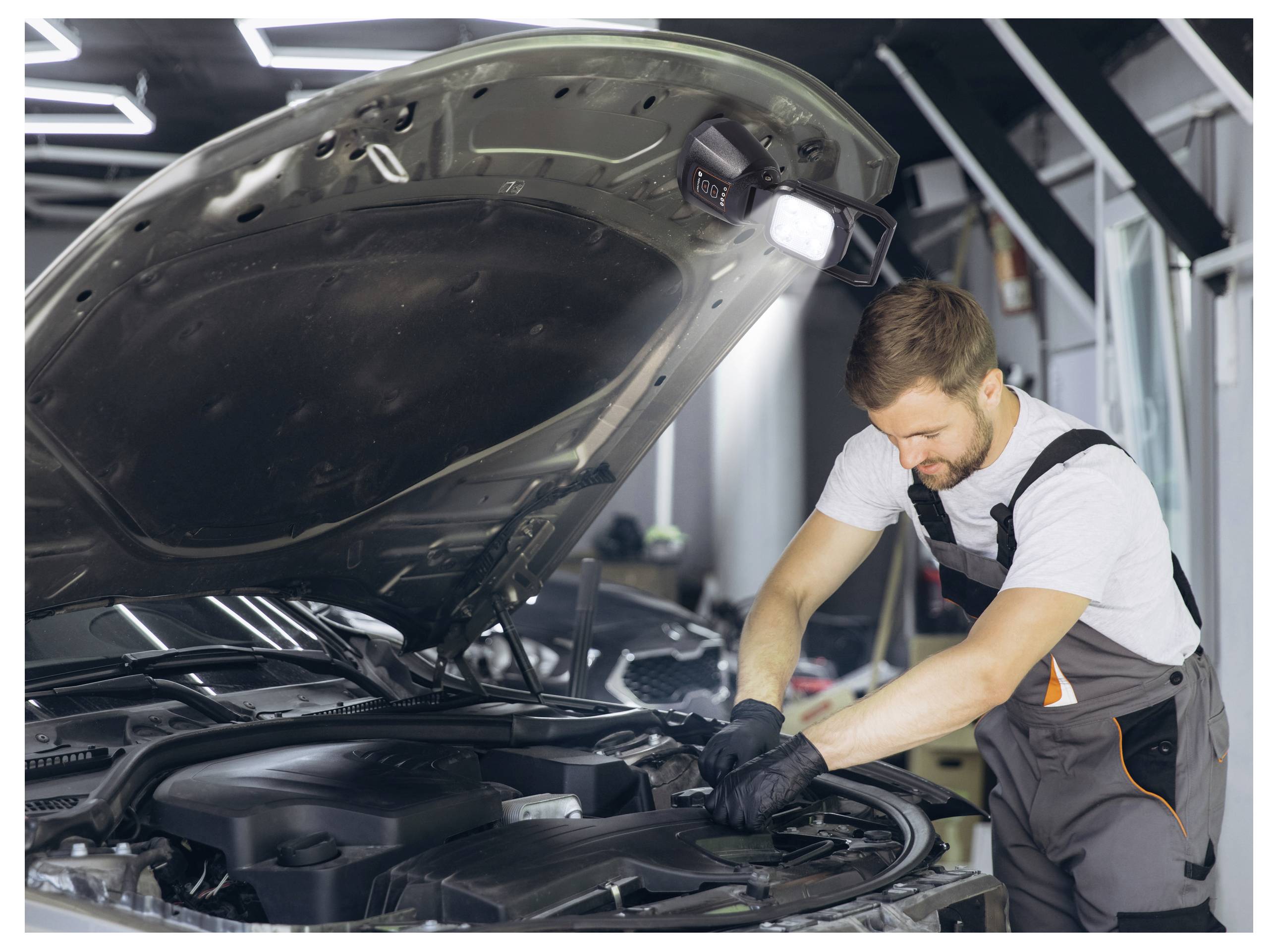 Een monteur in een garage werkt aan een automotor met de motorkap open, draagt werkhandschoenen en overall, gefocust op een reparatieklus.