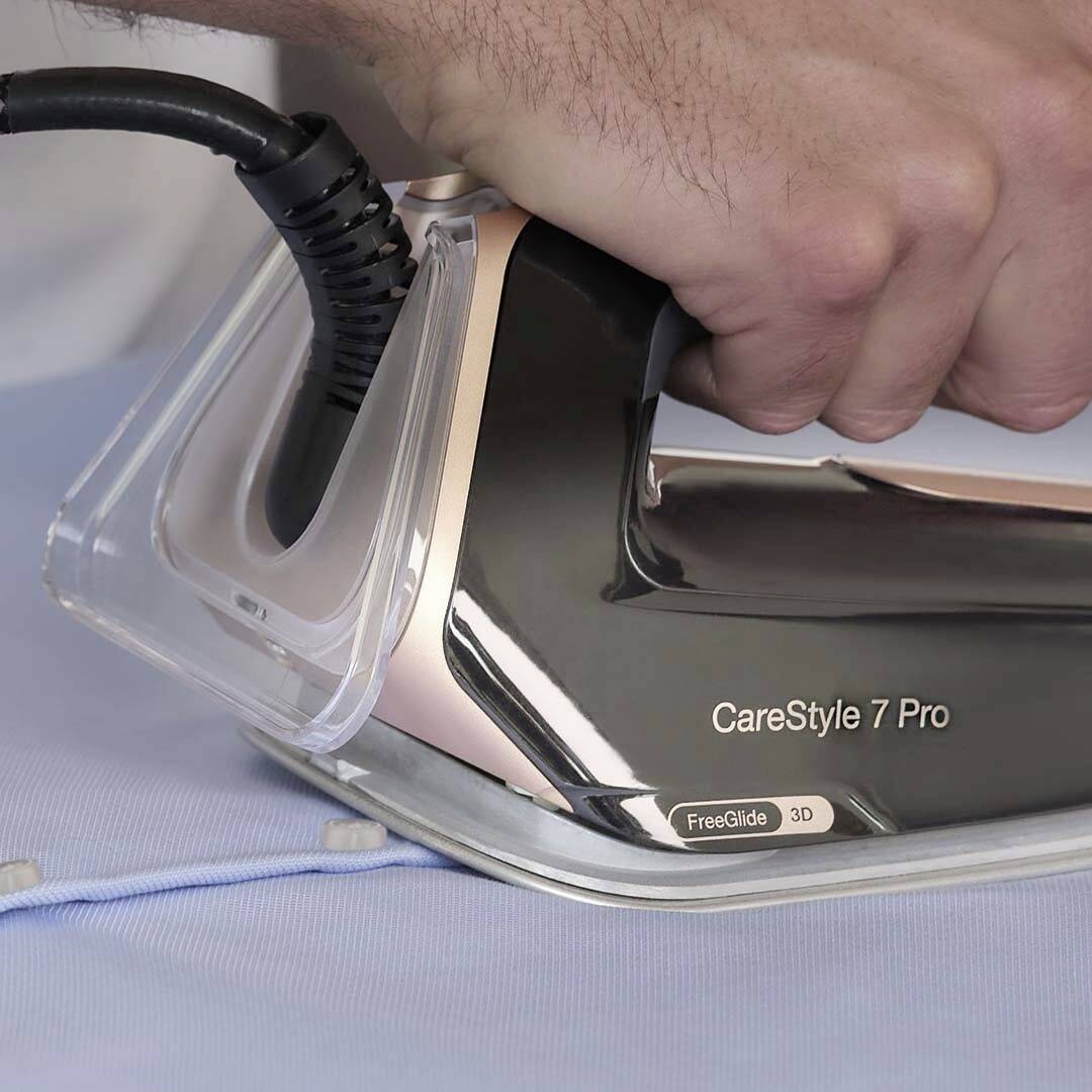 Een persoon die een zwarte en koperen stoomstrijkijzer met het label 'CareStyle 7 Pro' gebruikt op een lichtblauwe kledingstuk, waarbij het hoekige ontwerp van de strijkijzer wordt getoond.