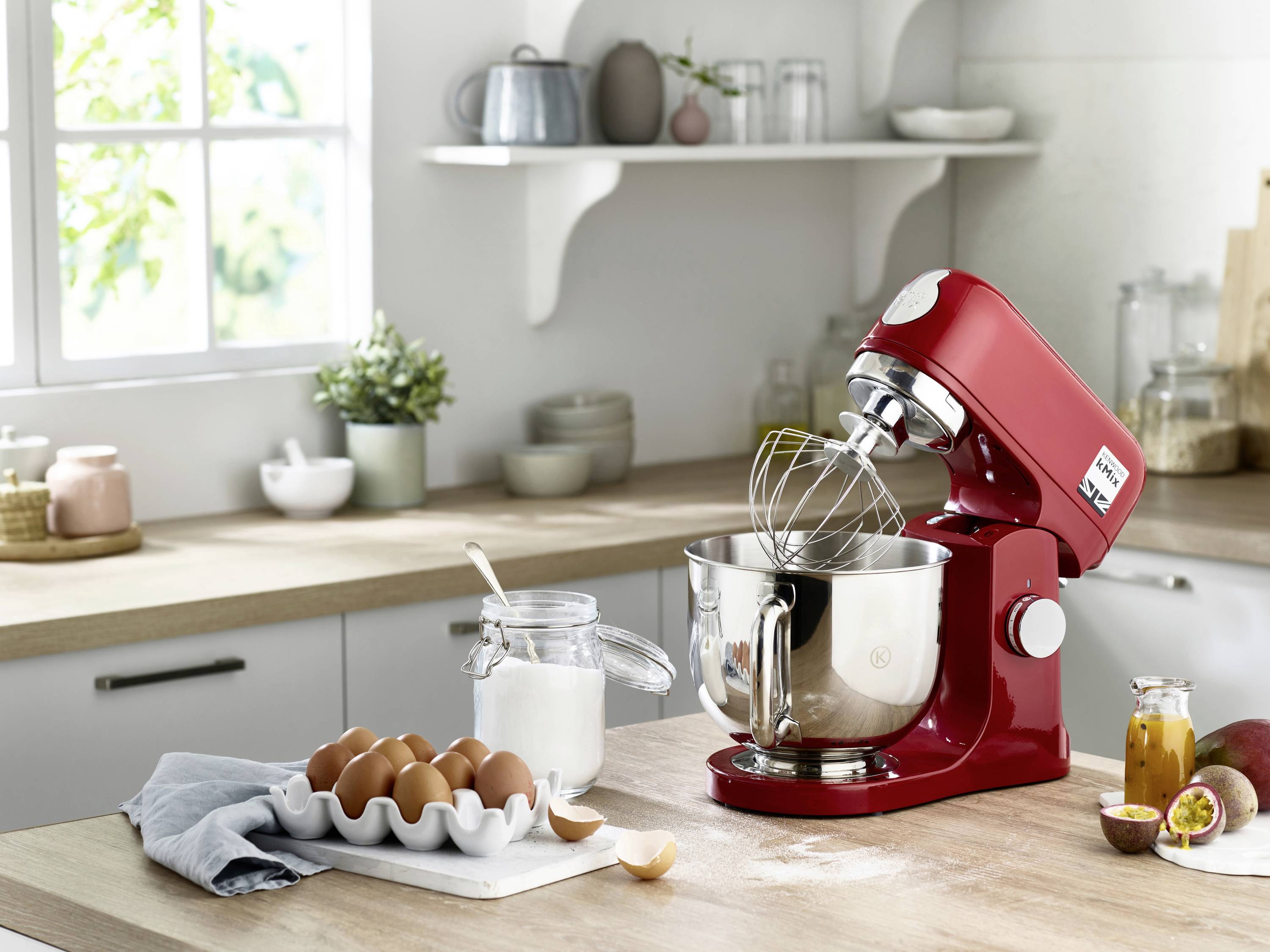 Een rode standmixer op een aanrecht met een klopper bevestigd in een metalen kom. Nabij staan een pot suiker en een pak eieren.