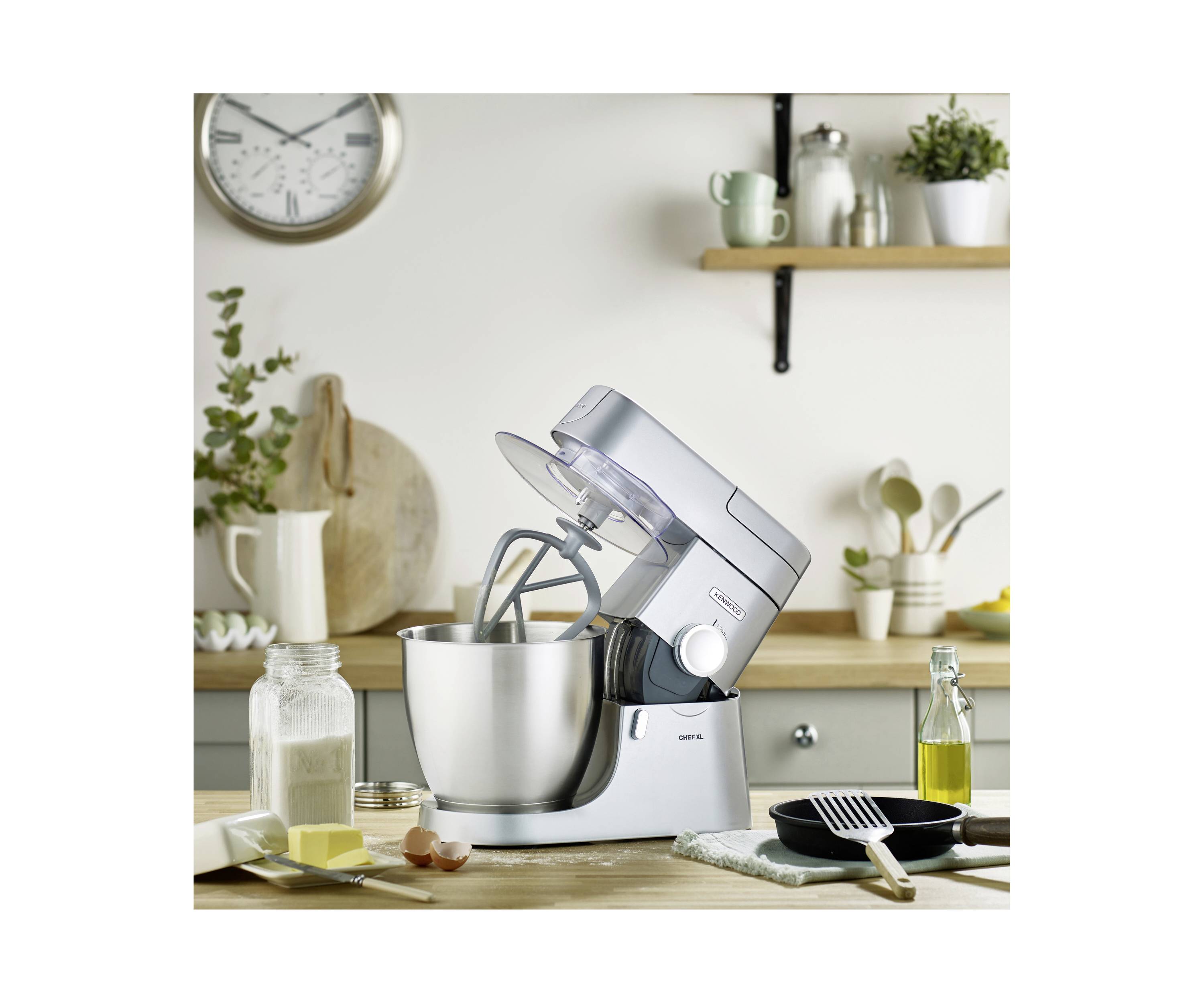 Een zilveren standmixer op een aanrecht met bakingredïenten, keukengerei en een wandklok op de achtergrond.