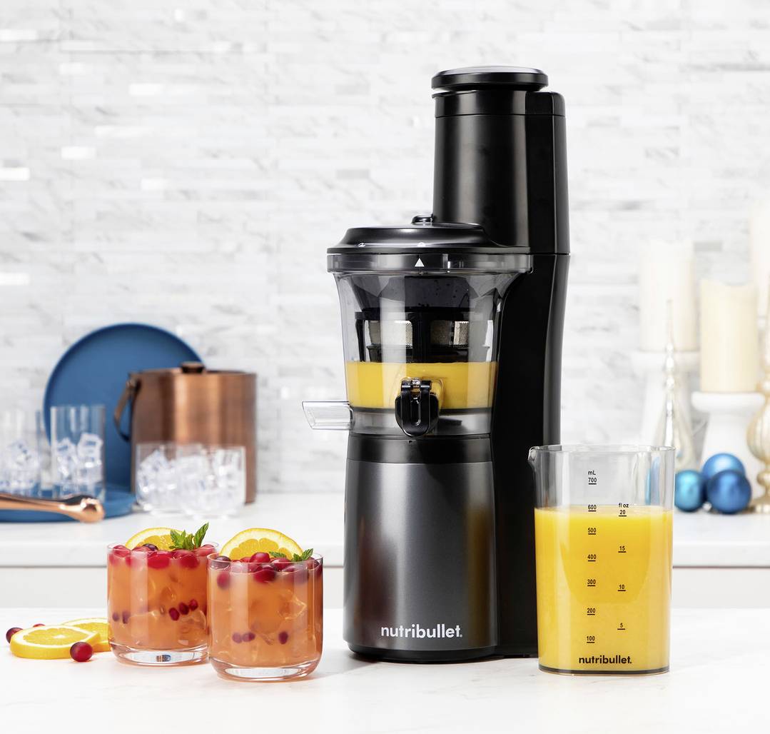 Een zwarte Nutribullet slowjuicer op een keukentafel met twee glazen kleurrijke fruitsap en een kan gevuld met sinaasappelsap ernaast.