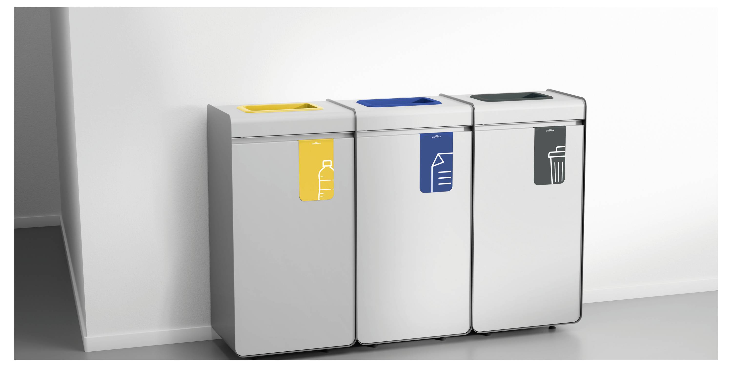 Drie afvalbakken voor recycling: geel voor flessen, blauw voor papier, en grijs voor restafval, tegen een witte muur binnenshuis.