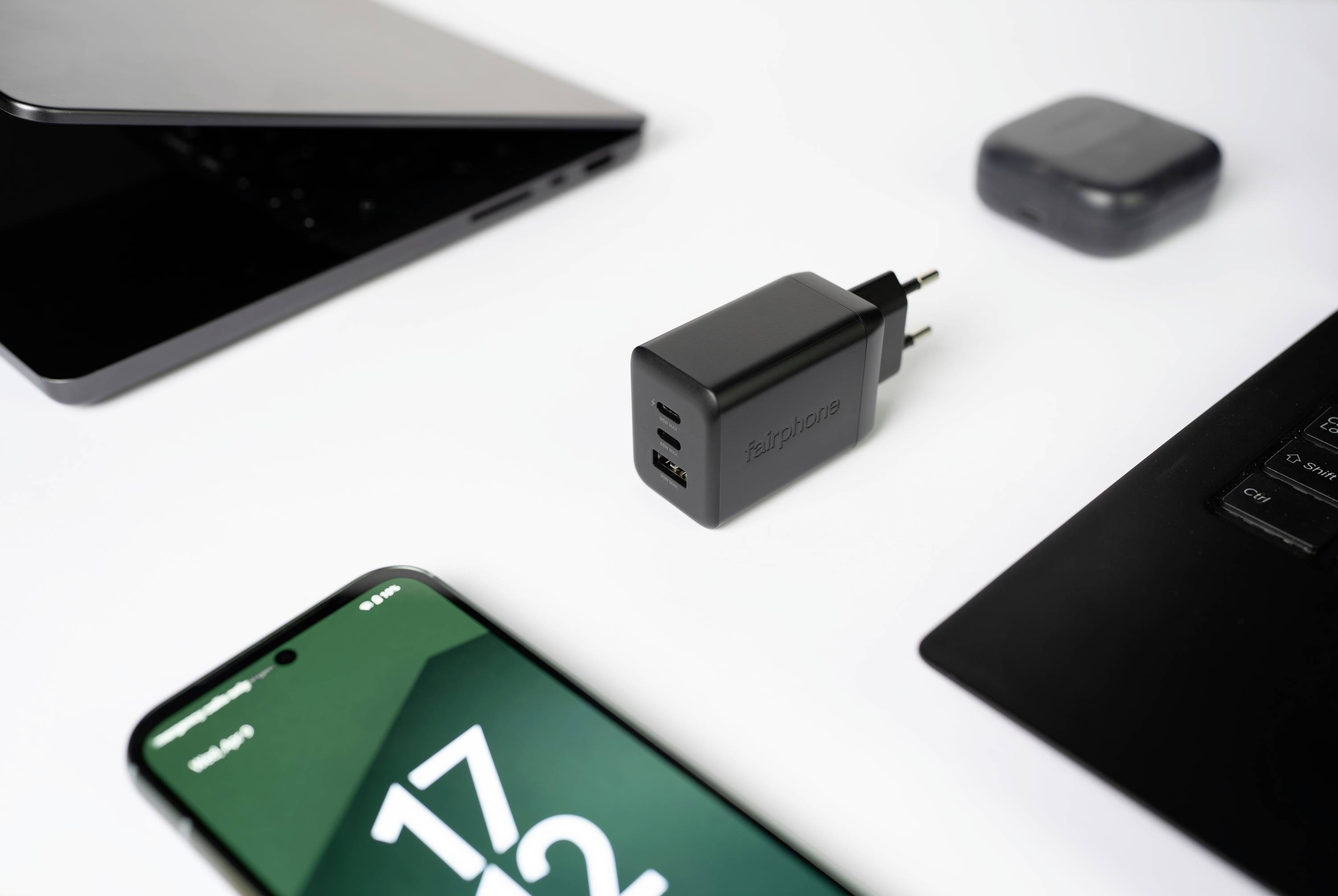 Een zwarte twee-poorts USB-wandoplader geplaatst tussen een smartphone met '17:23' op het scherm, een gedeeltelijk opengeslagen laptop en draadloze oordopjes op een wit oppervlak.
