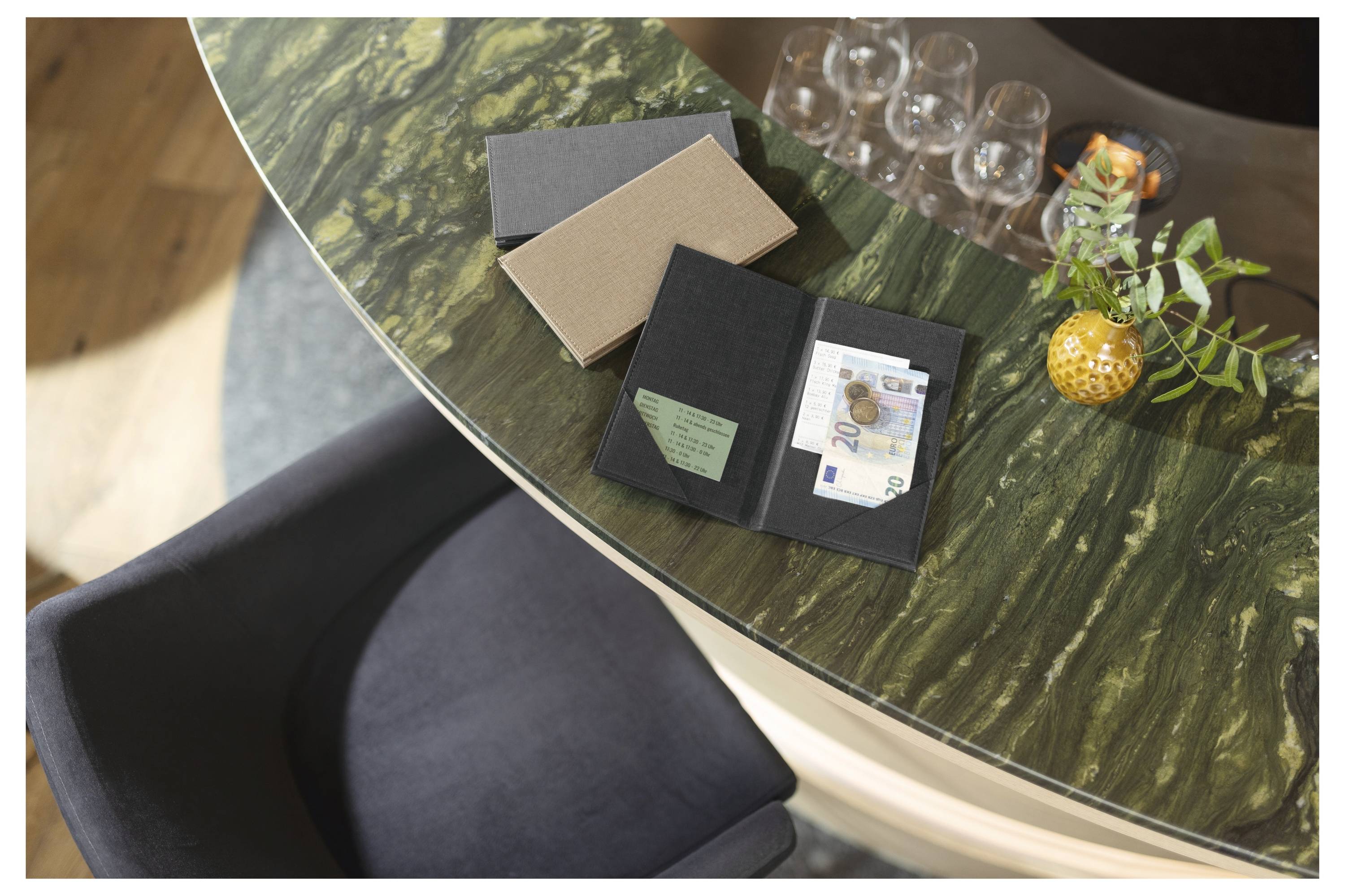 Open zwart portemonnee op een groen marmeren tafel met contant geld, kaarten, twee gesloten notitieboeken, bril en een klein plantje als tafelstuk.