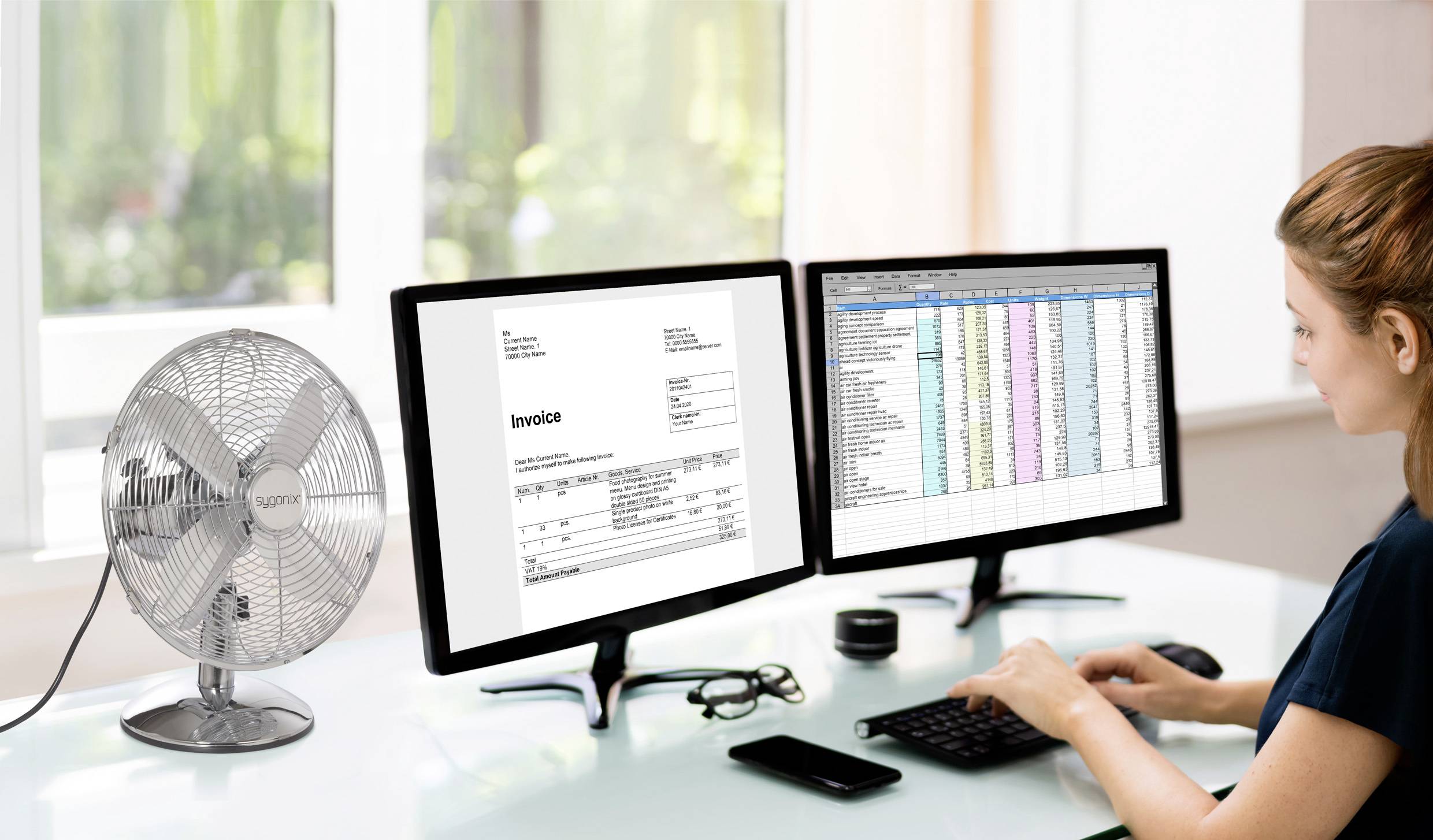 Een persoon werkt aan een bureau met twee computermonitors waarop een factuur en een spreadsheet worden weergegeven, naast een zilveren bureauventilator.