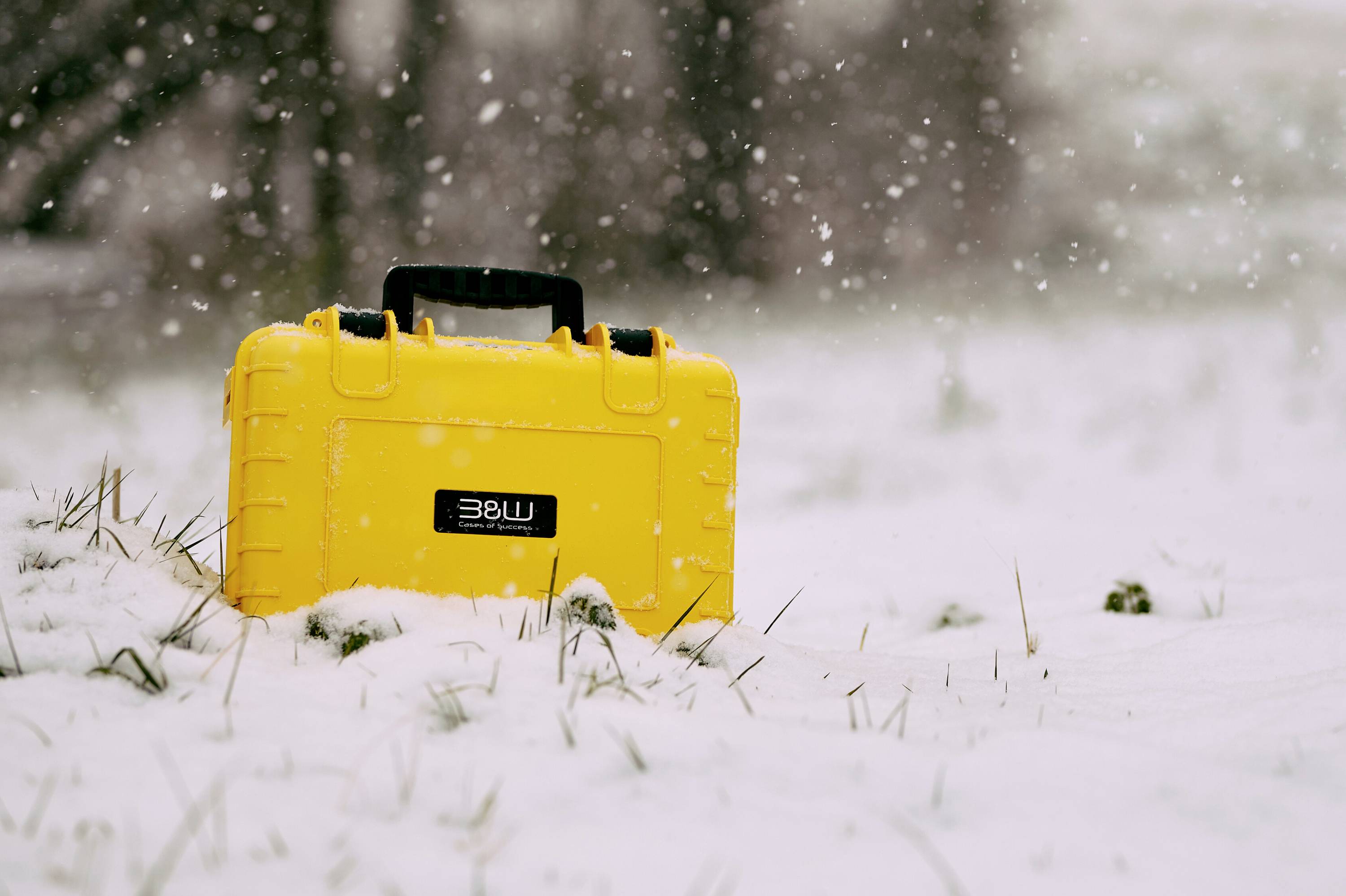 Een felgele beschermende behuizing met een handgreep die gedeeltelijk bedolven is onder de sneeuw, geplaatst in een besneeuwde buitenomgeving met vallende sneeuwvlokken.