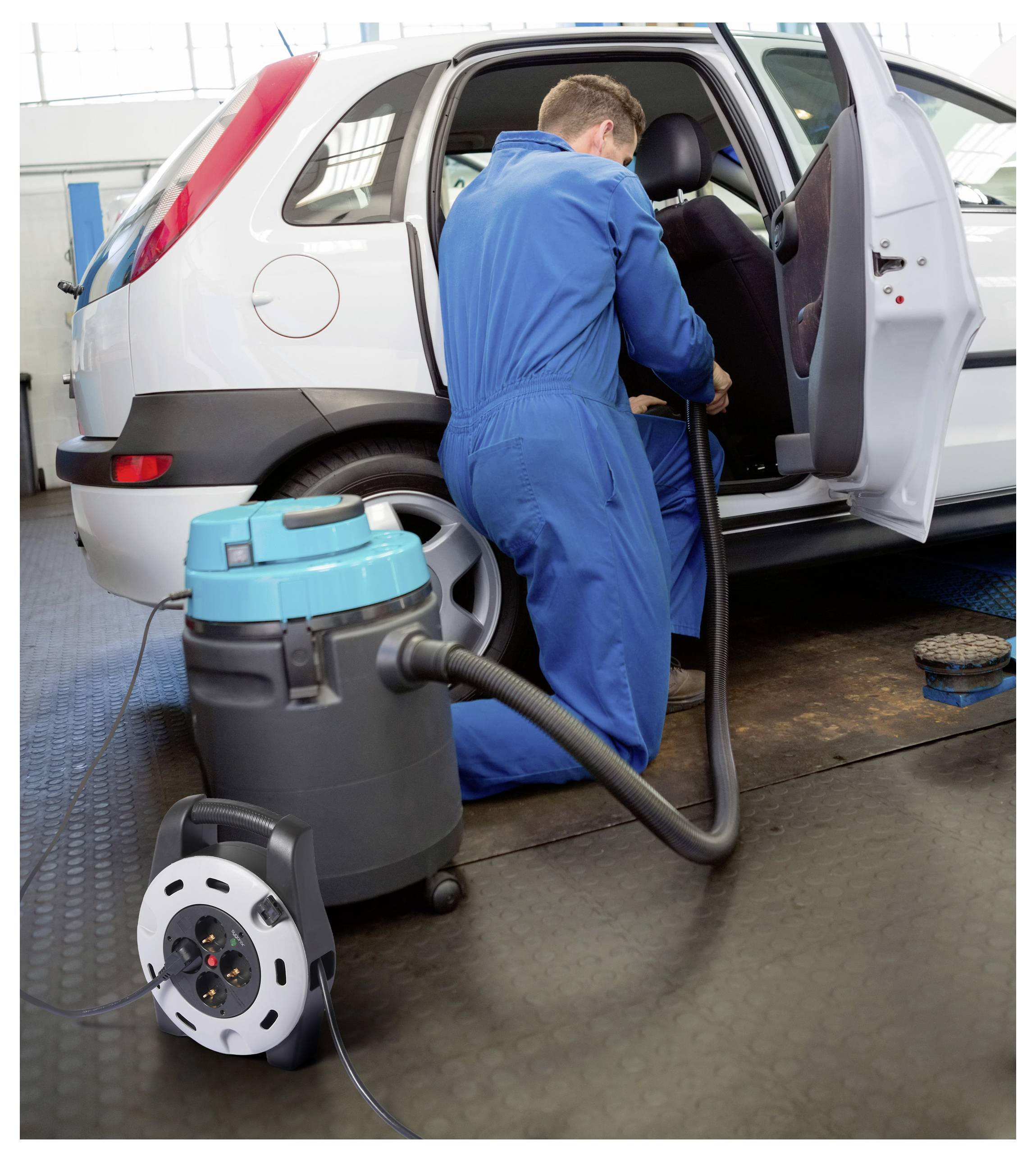 Een werknemer in blauwe overalls gebruikt een stofzuiger om het interieur van een witte auto in een garage schoon te maken. Een draagbare stofzuiger staat op de grond.