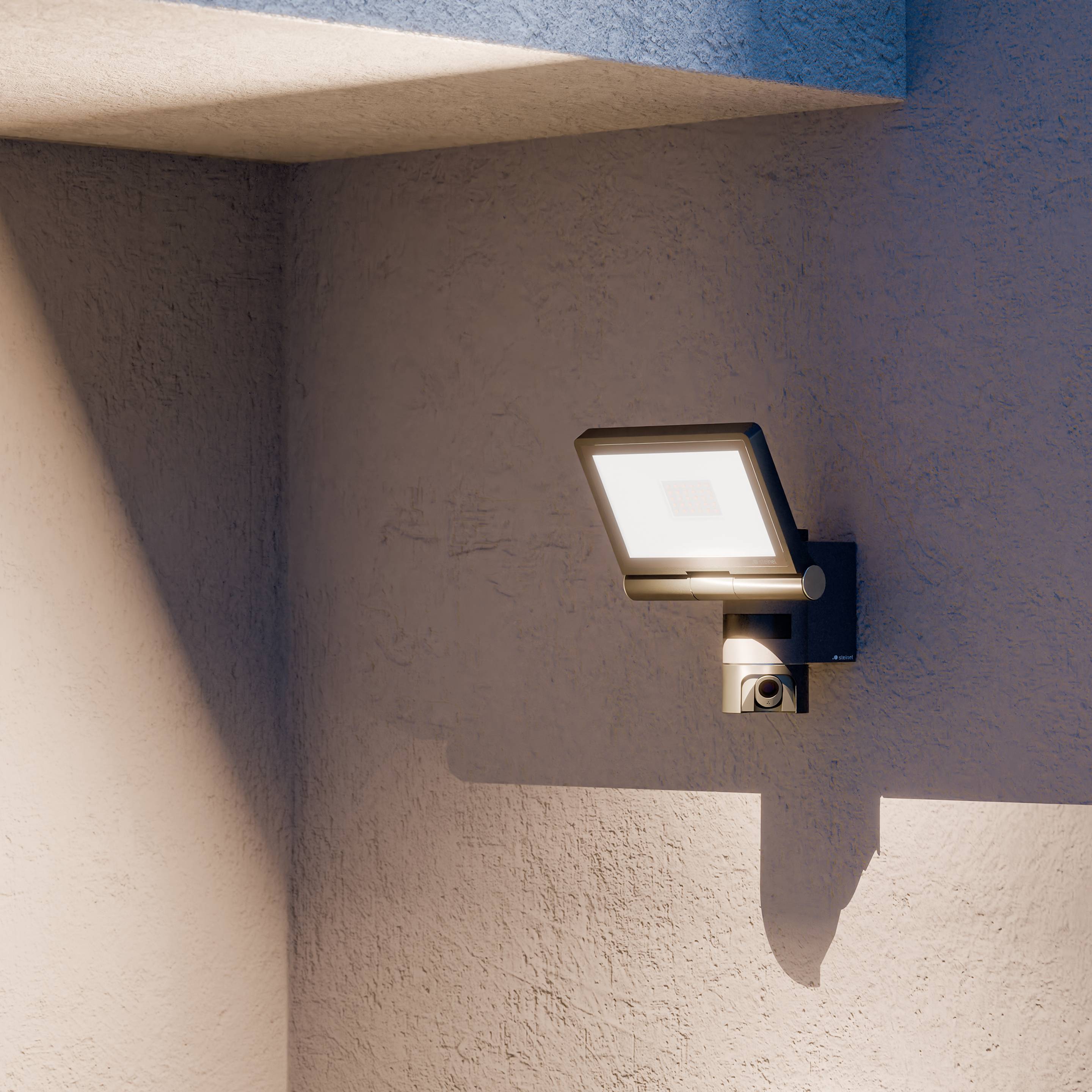 Een aan de muur bevestigde LED-lamp 's avonds, die een geometrische schaduw werpt op een pleistermuur onder een moderne betonnen uitbouw, waardoor een serene sfeer ontstaat.