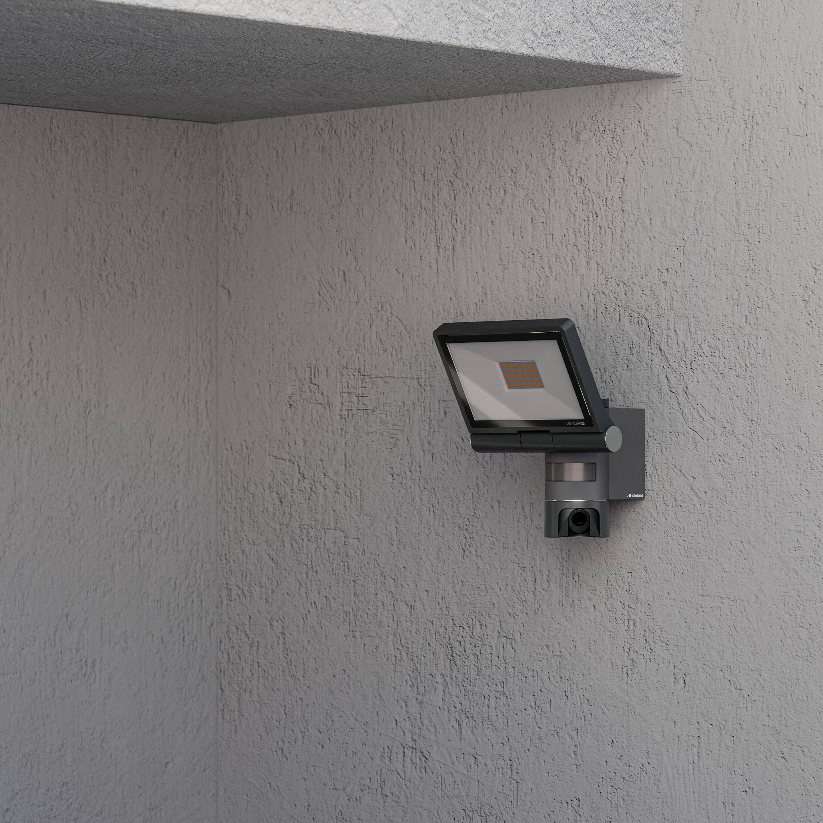 Een aan de muur bevestigde buitenbeveiligingslamp met bewegingssensor, bevestigd aan een grijze betonnen muur. Hij is geplaatst onder een uitstekende rand van een gebouw.