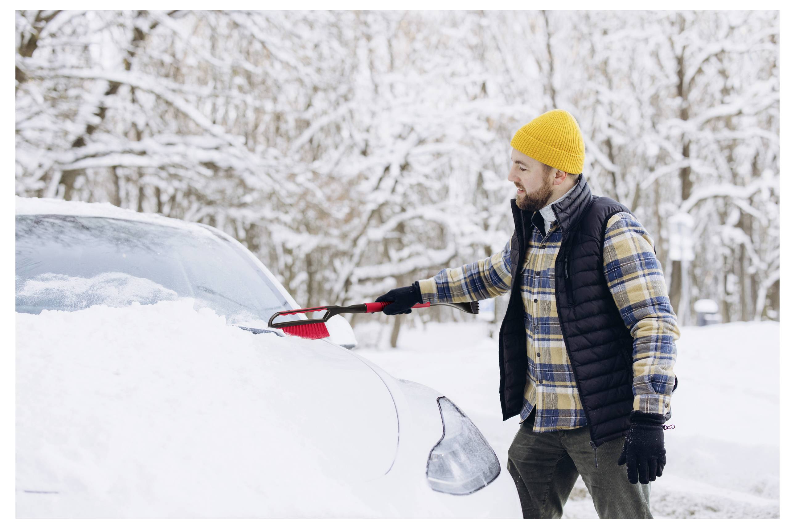 Een persoon in een gele hoed en geruit jasje gebruikt een sneeuwborstel om zware sneeuw van een auto af te vegen in een besneeuwde bosomgeving.