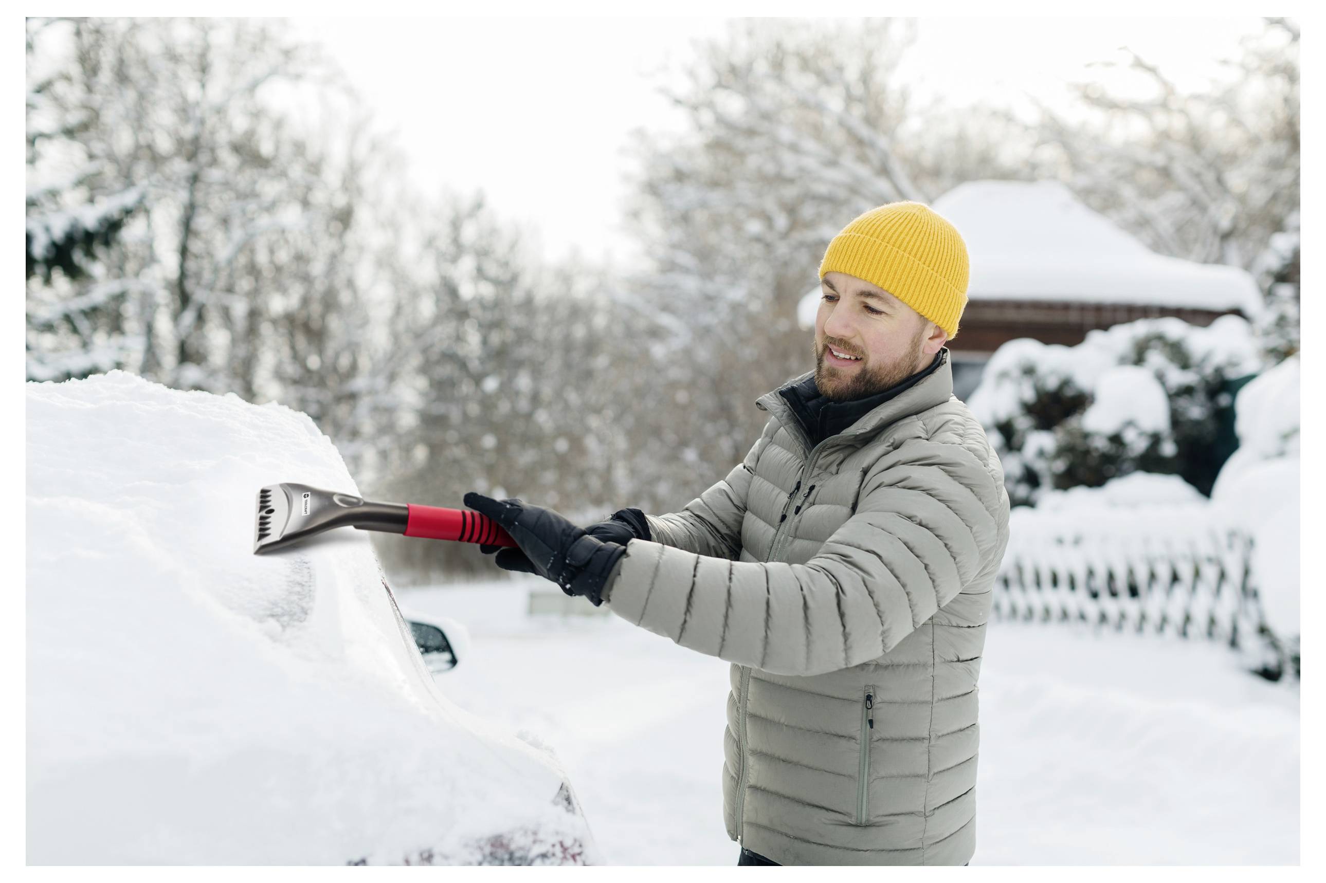 Man met een gele muts en grijze jas verwijdert sneeuw van een auto met een ijskrabber in een sneeuwrijke woonwijk.