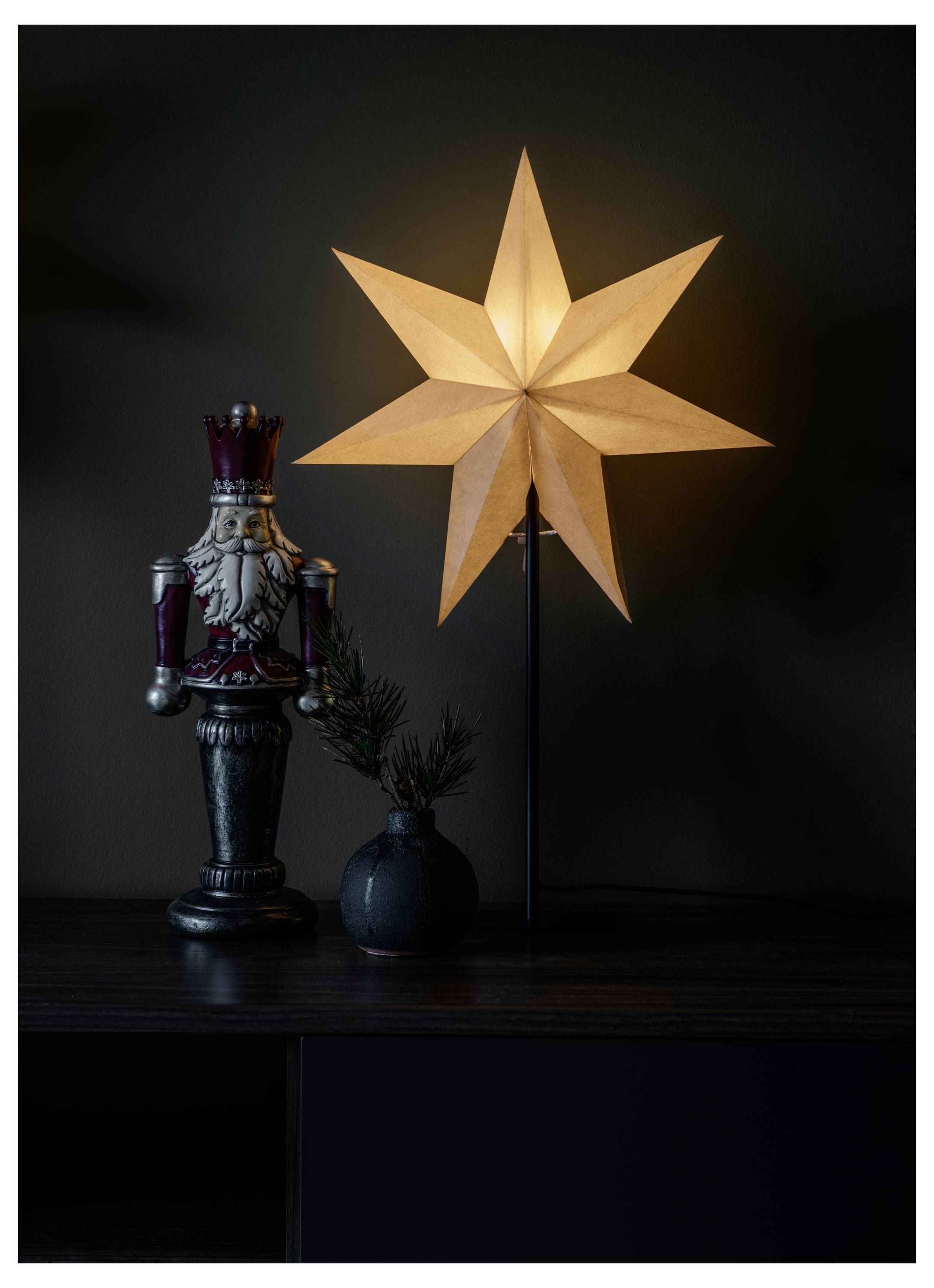 Een gloeiende stervormige lamp op een standaard naast een decoratieve notenkraker-figuur en een kleine vaas met dennentakken op een donker oppervlak.