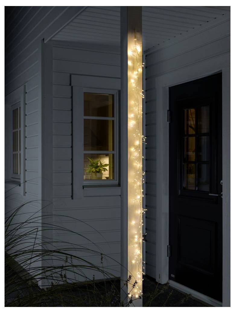 Een zuil op de veranda van een huis is omwikkeld met fonkelende kerstlichtjes. Vlakbij onthult een warm verlicht raam een potplant aan de binnenkant.