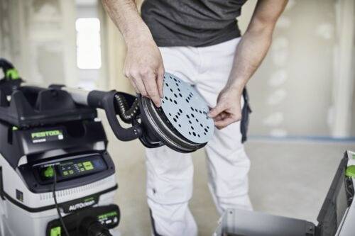 Een persoon stelt een schuurschijf af op een schuurmachine in een ruimte die wordt gerenoveerd met onafgewerkte muren.