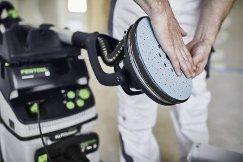 Een persoon stelt een schuurmachine af die is aangesloten op een stofafzuiger, en bereidt zich voor op het gladmaken van oppervlakken, wat wijst op een focus op oppervlakte-afwerkingswerkzaamheden.